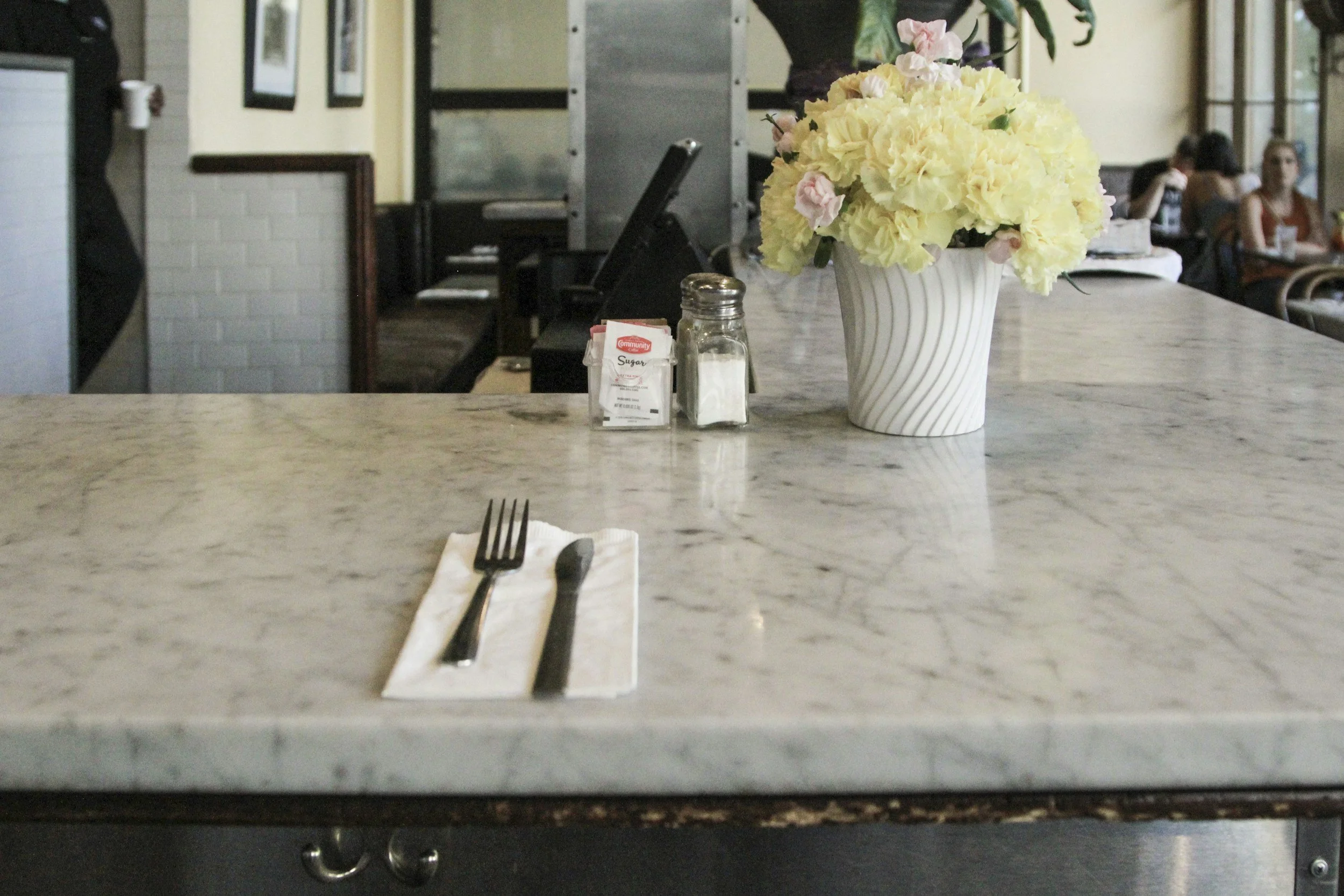 A marble-topped table in a restaurant with a fork and knife on a napkin, a glass jar of sugar, a sugar packet, and a large white vase filled with light yellow and pink flowers. In the background, there are other tables with people dining.
