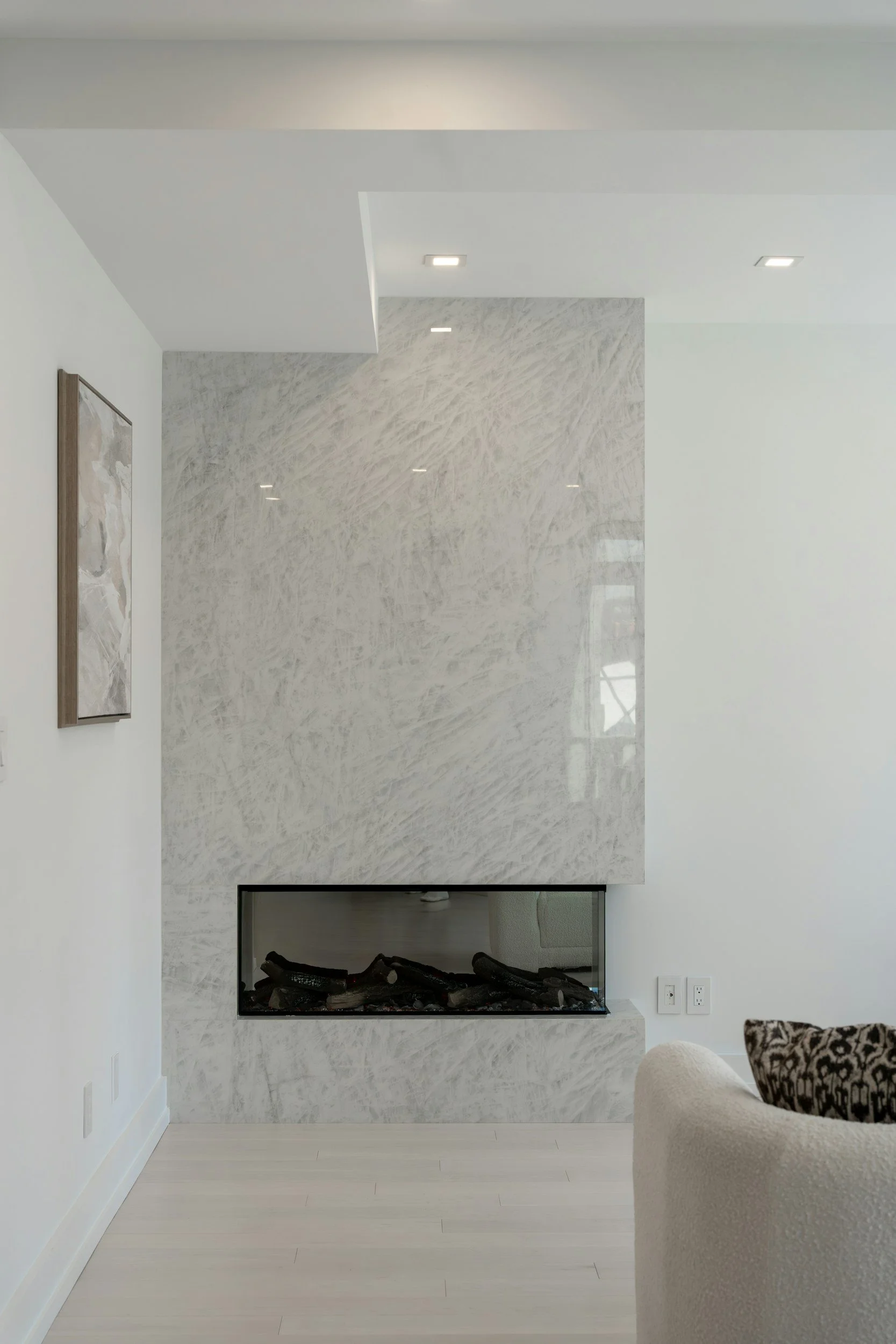 Modern living room with a white and gray fireplace, white walls, light-colored hardwood flooring, and a corner of an upholstered chair with a patterned pillow.