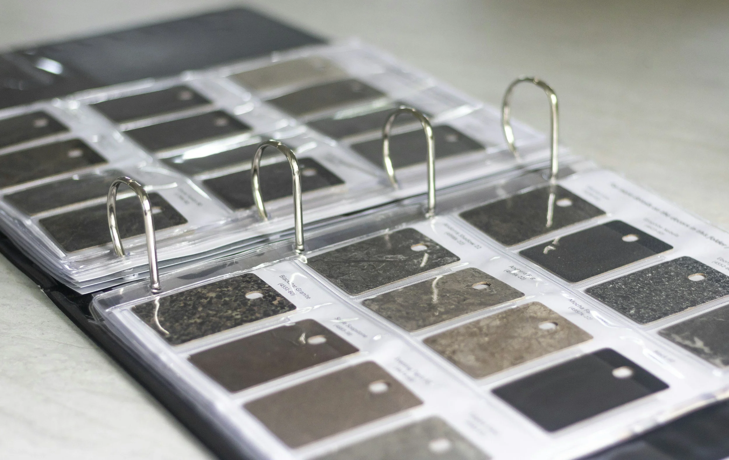 A binder containing multiple samples of dark-colored granite and marble slabs, each labeled with descriptions, placed on a table.