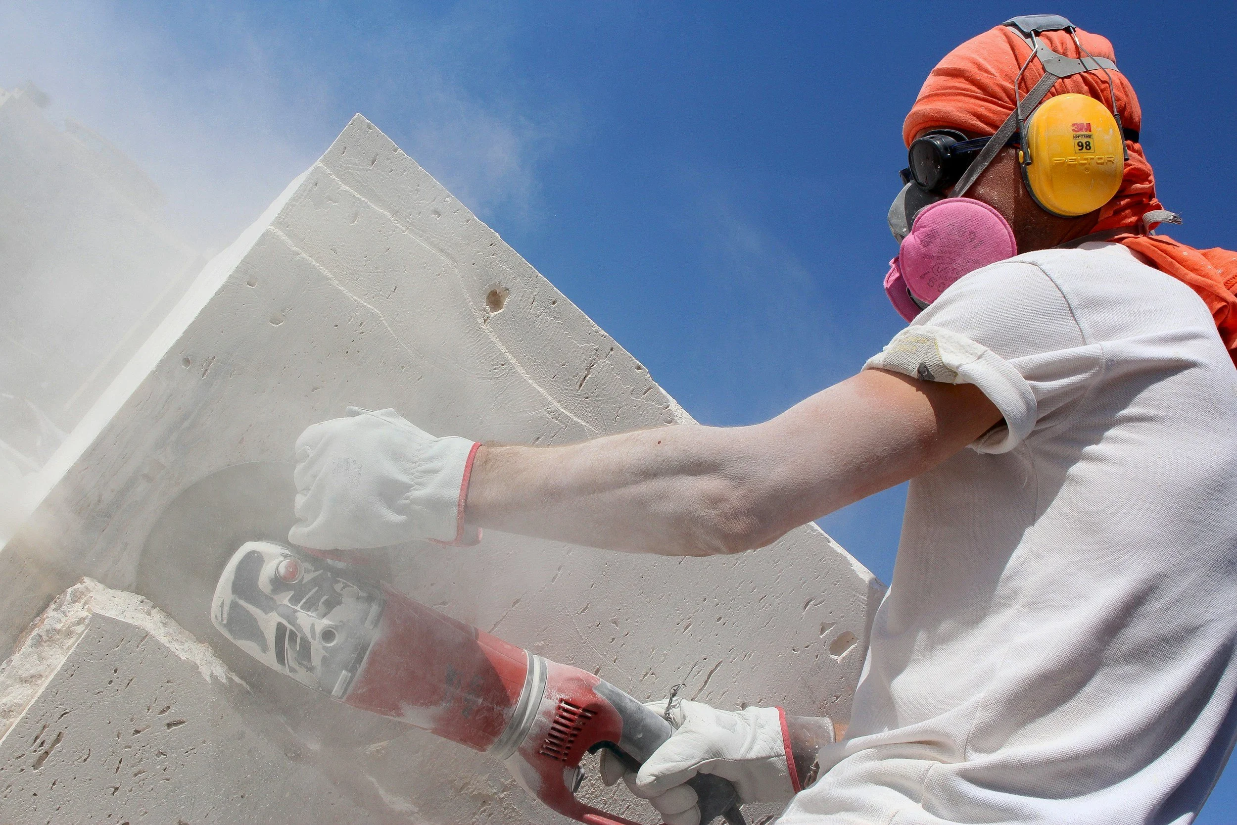 Person wearing a yellow ear protector, white shirt, pink face mask, and white gloves using a red power tool to cut white material against a blue sky background.