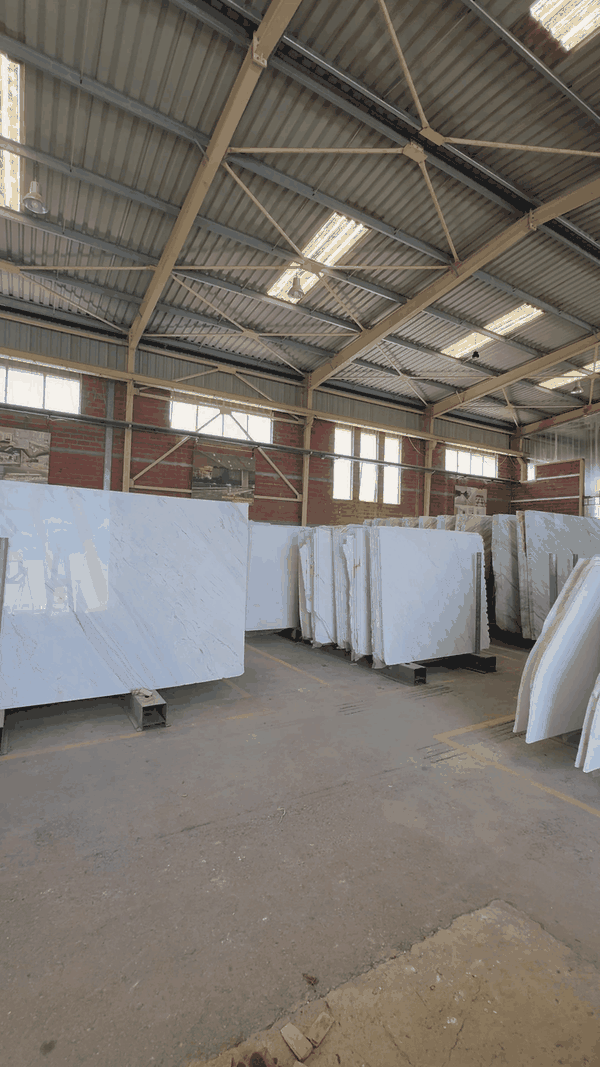Inside a warehouse with large white marble slabs stacked on black supports, with a metal roof and windows letting in natural light.