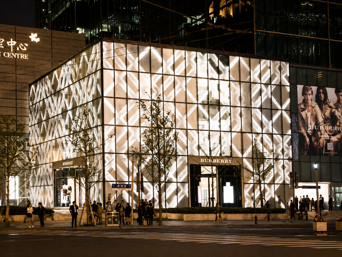 Exterior view of a modern Burberry store at night with illuminated geometric facade, trees, pedestrians, and a large advertisement featuring models wearing Burberry clothing.