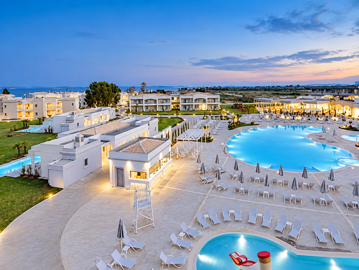 Twilight view of a resort with illuminated white buildings, swimming pools with lounge chairs, umbrellas, and cabanas, overlooking a scenic landscape and water body in the distance.