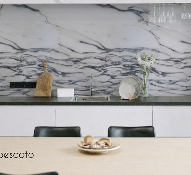 Modern kitchen with marble backsplash, green countertop, white lower cabinets, and a wooden dining table with a plate of bread rolls. There is a faucet and sink, a wooden cutting board, white soap or sponge, and a glass vase with white flowers on the countertop. The table has black chairs.