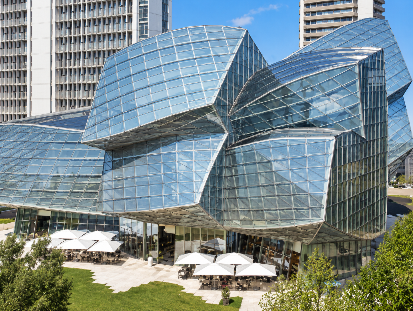 Modern glass building with irregular geometric shapes, surrounded by greenery and outdoor seating with white umbrellas, in an urban setting with tall buildings in the background.