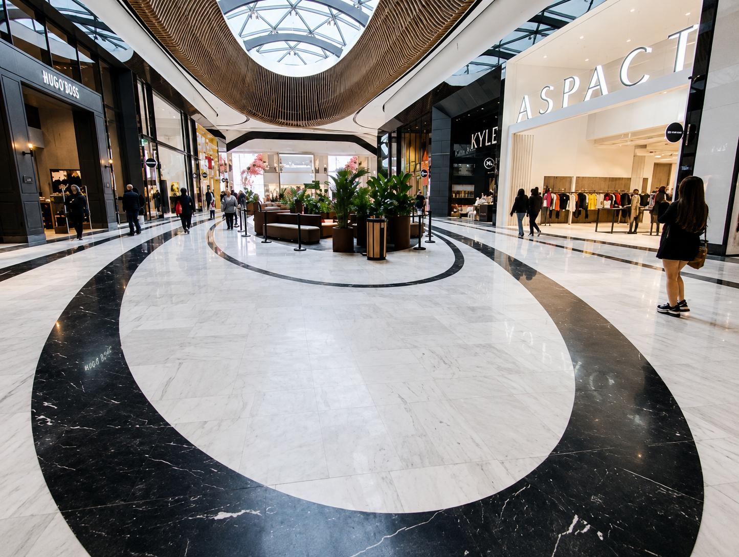 An indoor shopping mall with a marble floor featuring black and white decorative circles. Several people are walking, and storefronts for Hugo Boss, Aspaci, and Kyle are visible on either side. There are potted plants and a seating area in the center. The ceiling has a glass dome allowing natural light.