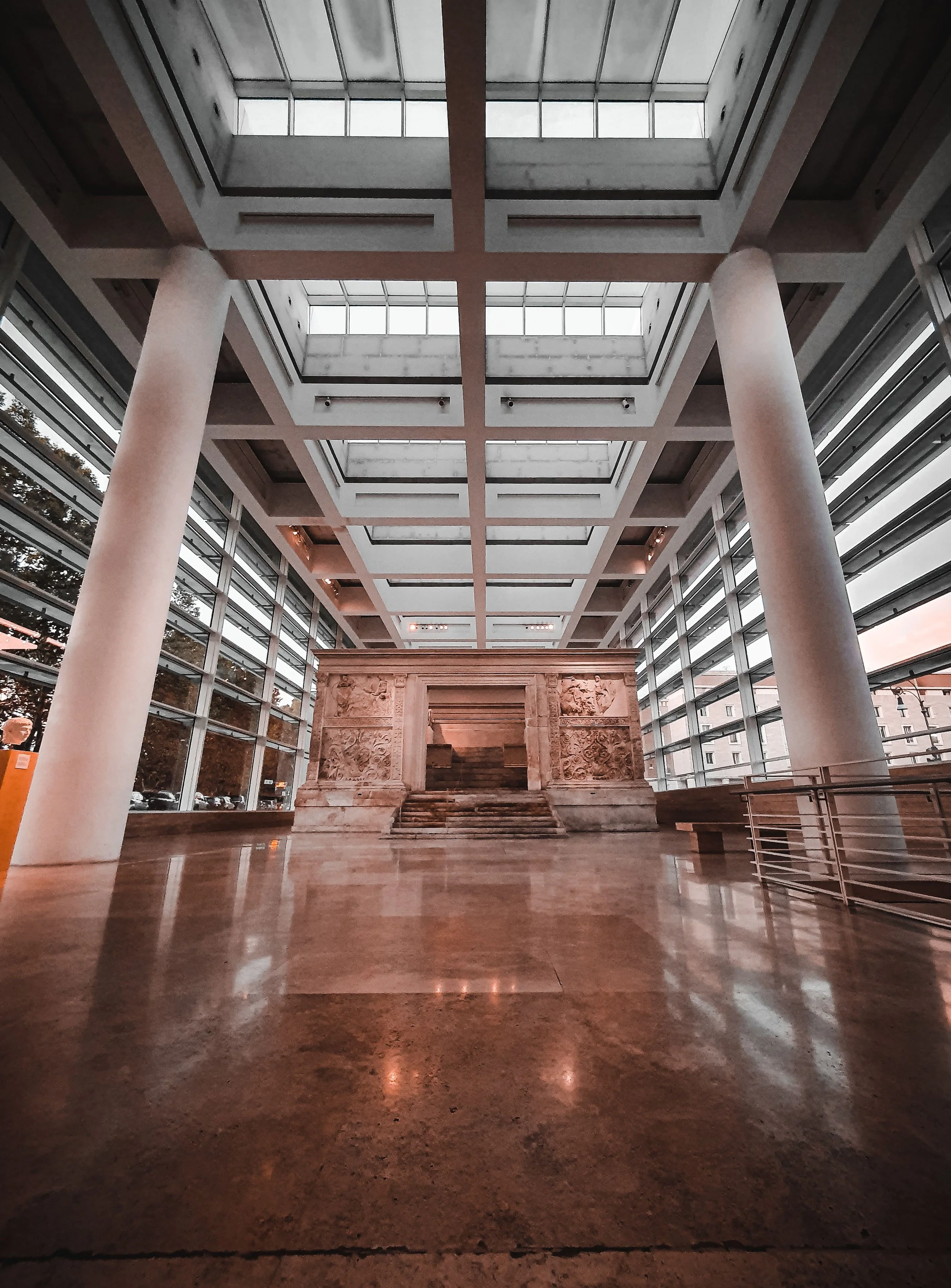 Interior of a modern building with a high ceiling, large marble columns, and a carved stone staircase at the center. Skylights provide natural light, and glass walls with metal framing surround the space.