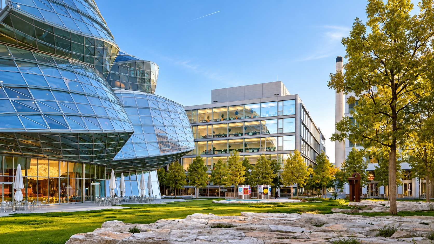 Modern glass building with outdoor seating area, green trees, and a grassy landscape under a clear blue sky.