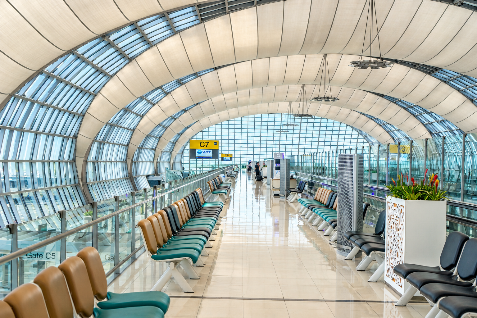 Empty airport terminal with many chairs, large glass windows, and a few passengers with luggage, signs for gates C7 and C5, and a flower arrangement.