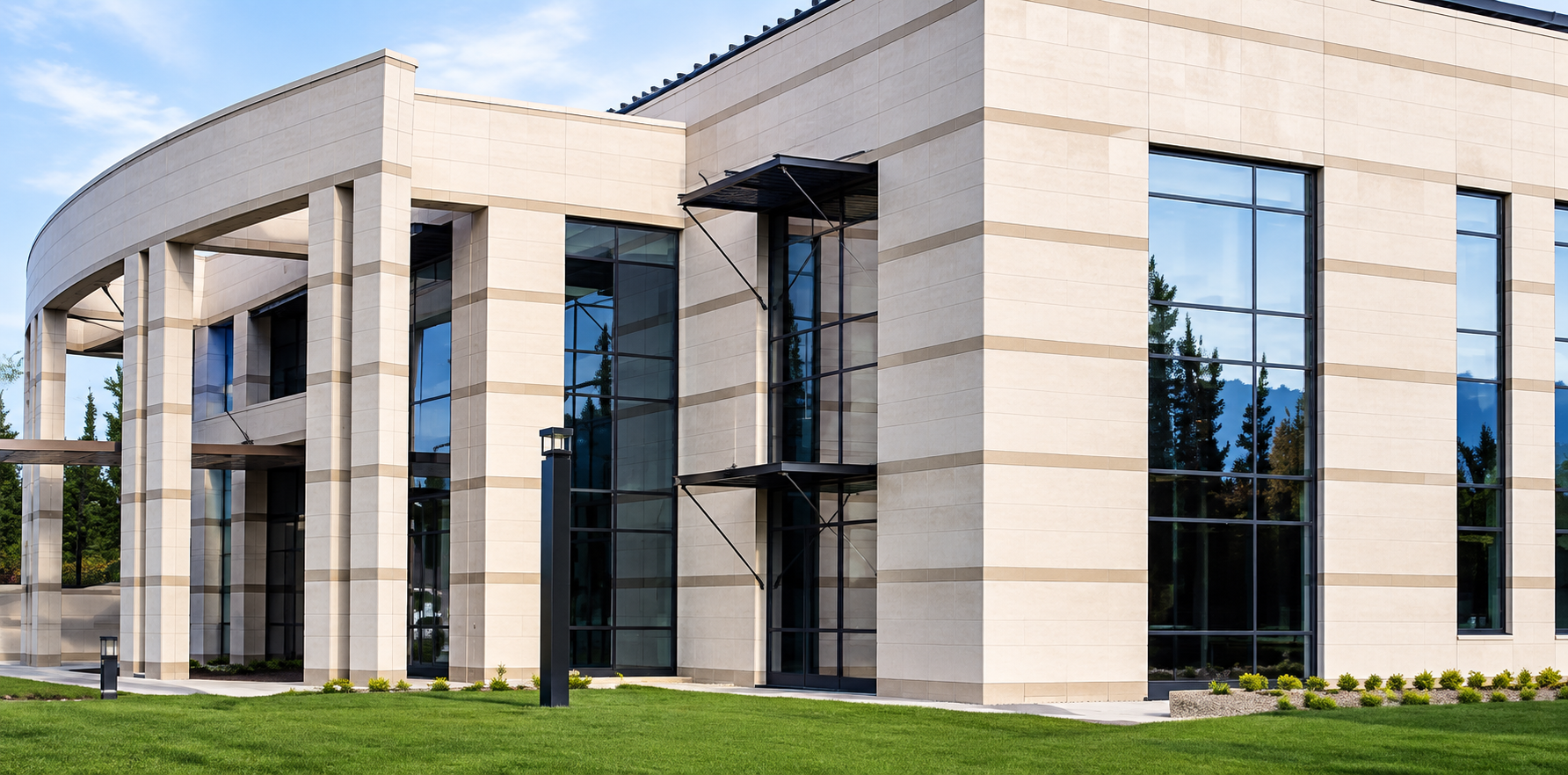 Modern commercial building with large glass windows and beige stone facade, surrounded by a well-manicured lawn and trees in the background.