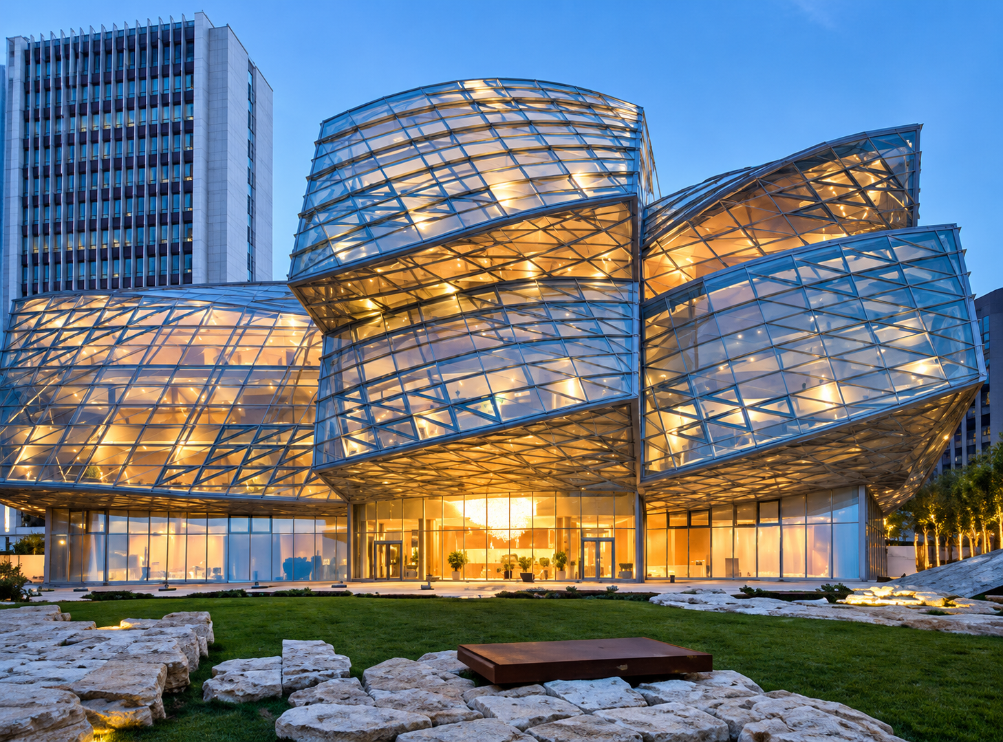 Modern building with a glass and metal facade illuminated from within at dusk, surrounded by landscaped rocks and greenery.