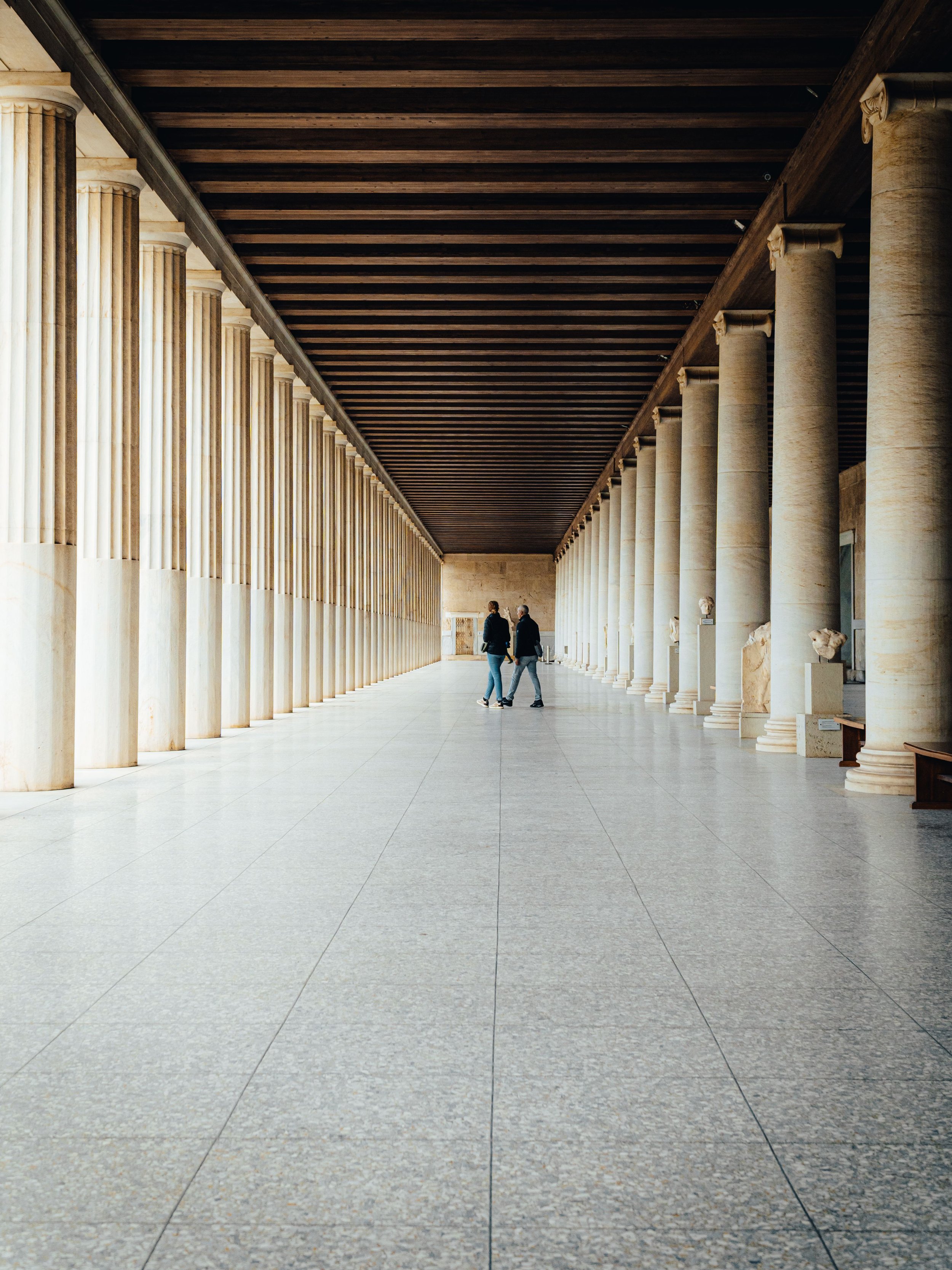 View of ancient Greek-style columns and statues inside a building, with two people walking and talking in the distance.