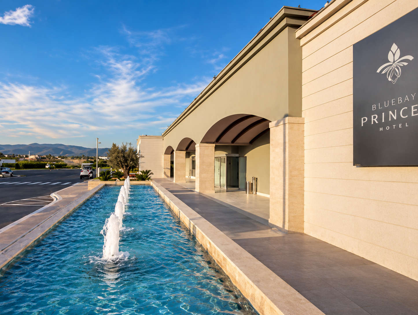 Exterior view of the Bluebay Princess Hotel showing a water fountain with multiple jets along a walkway in front of a beige building with arches, under a blue sky with scattered clouds and mountains in the background.