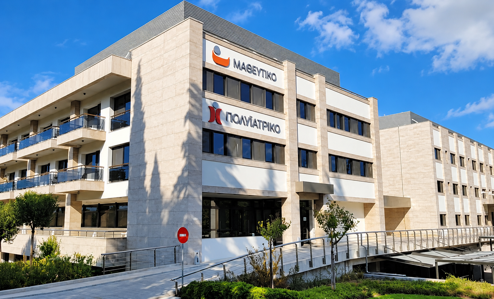 Modern multi-story medical building with signs in Greek for Magnetiko and Poliatreio, surrounded by small trees, bushes, and a bright blue sky.