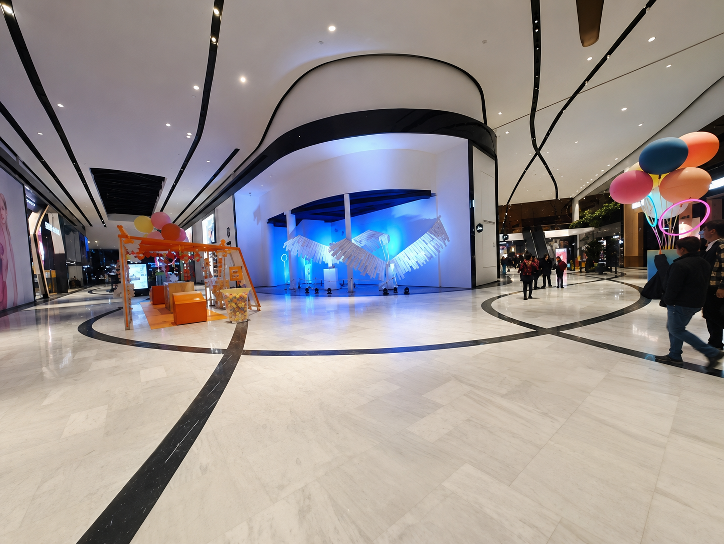 Indoor shopping mall decorated with colorful balloons and light displays, including a blue piano sculpture and an orange swing set with a small seating area.