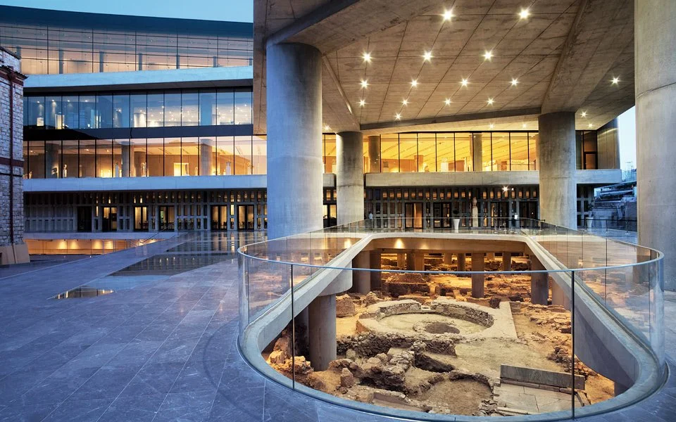 Modern building with large glass windows and a curved balcony, showcasing an archaeological excavation site in the foreground.