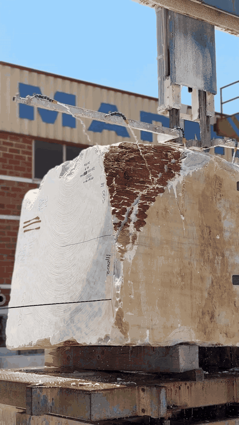 Large block of raw marble stone with rough cut surface on a wooden pallet outdoors under a clear blue sky, near a brick building.