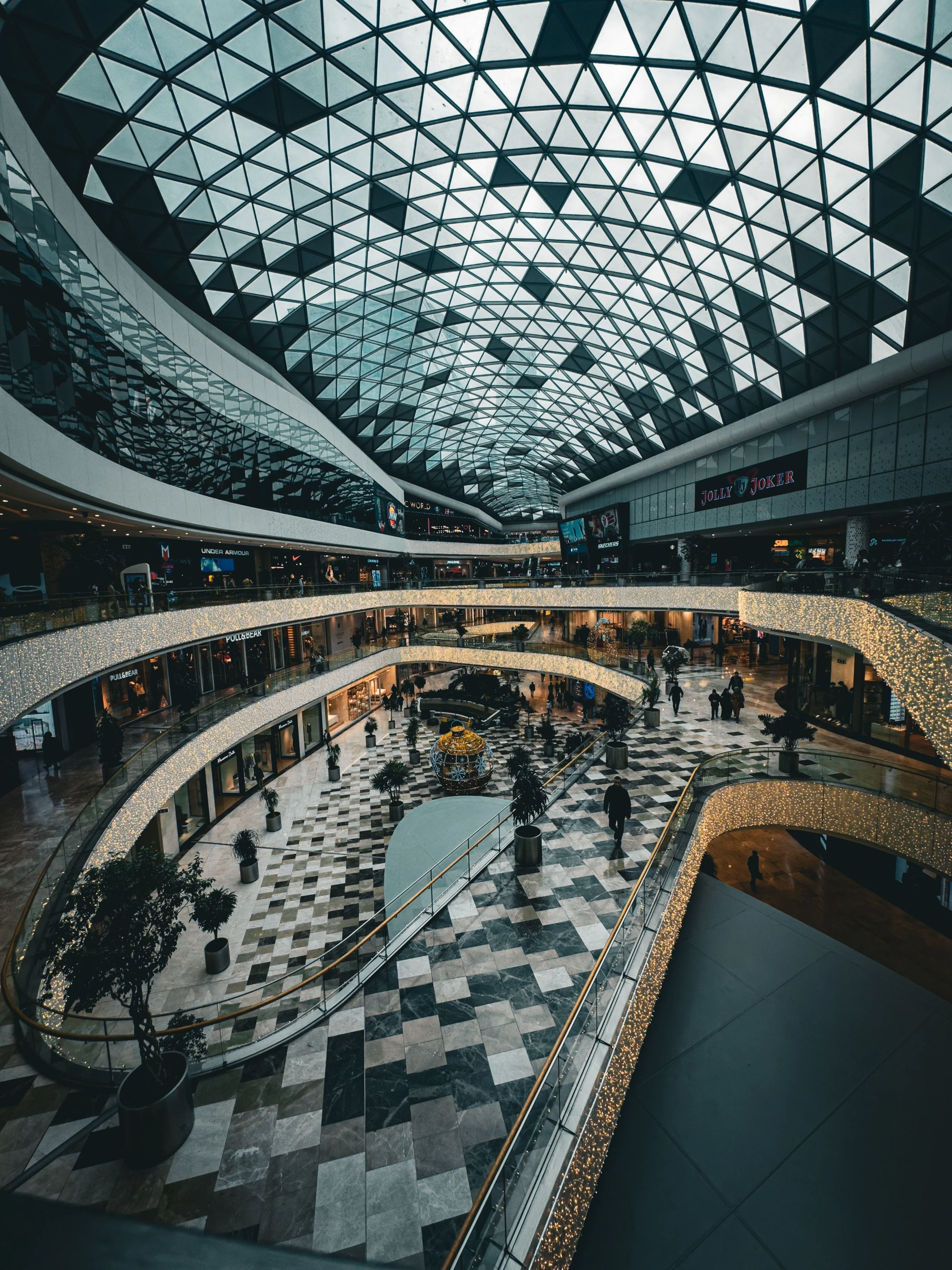 Interior view of a modern multi-level shopping mall with a glass, geometrically-patterned roof and busy walkways filled with shoppers. Stores and signs are visible on each level.