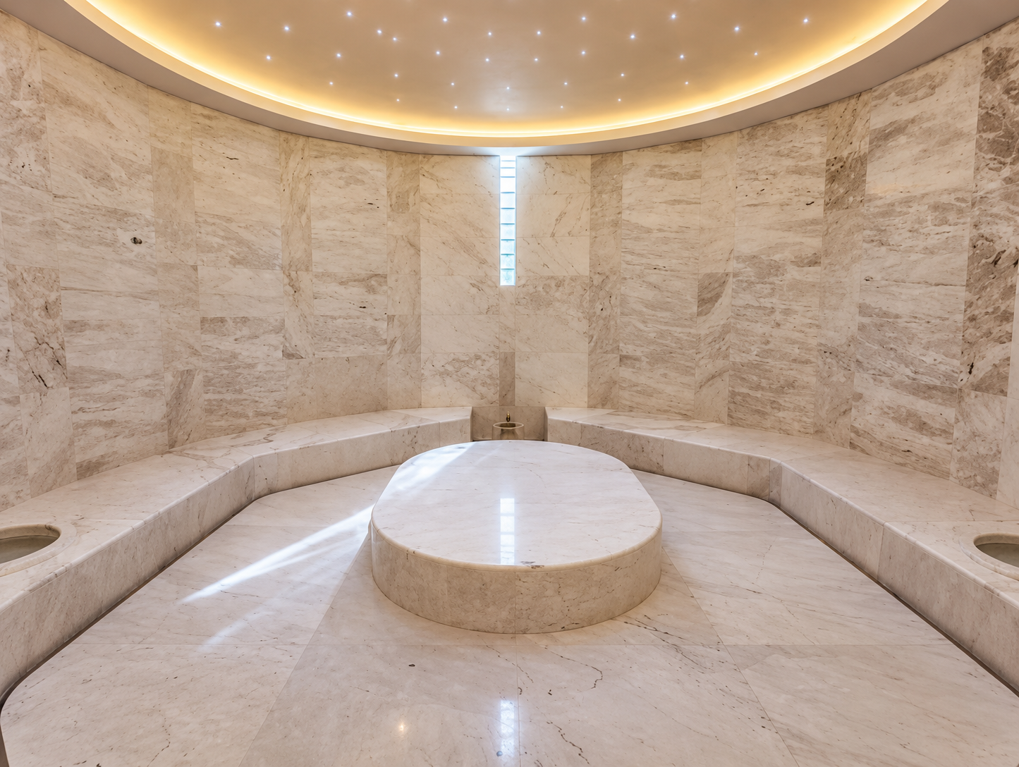 A spacious, circular marble room with built-in seating along the walls, a central raised marble platform, and a small window with glass blocks letting in light. The ceiling has recessed lighting and a soft glow around the edges.