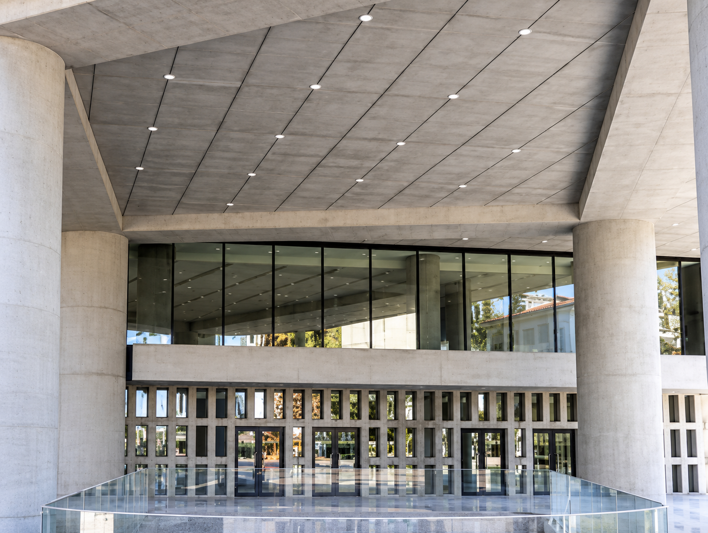 Modern architectural building with large glass windows, concrete columns, and a sloped concrete ceiling with recessed lighting.
