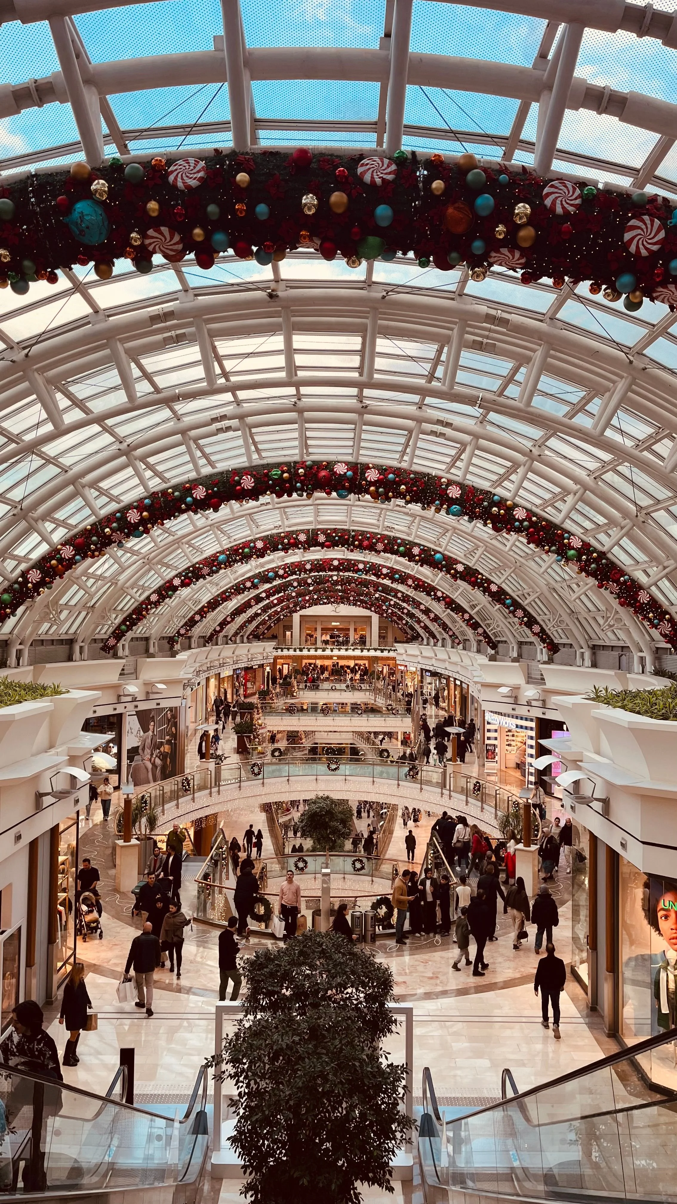 Interior view of a shopping mall decorated with Christmas ornaments and garlands hanging from the arched glass ceiling, with shoppers walking around on multiple levels.