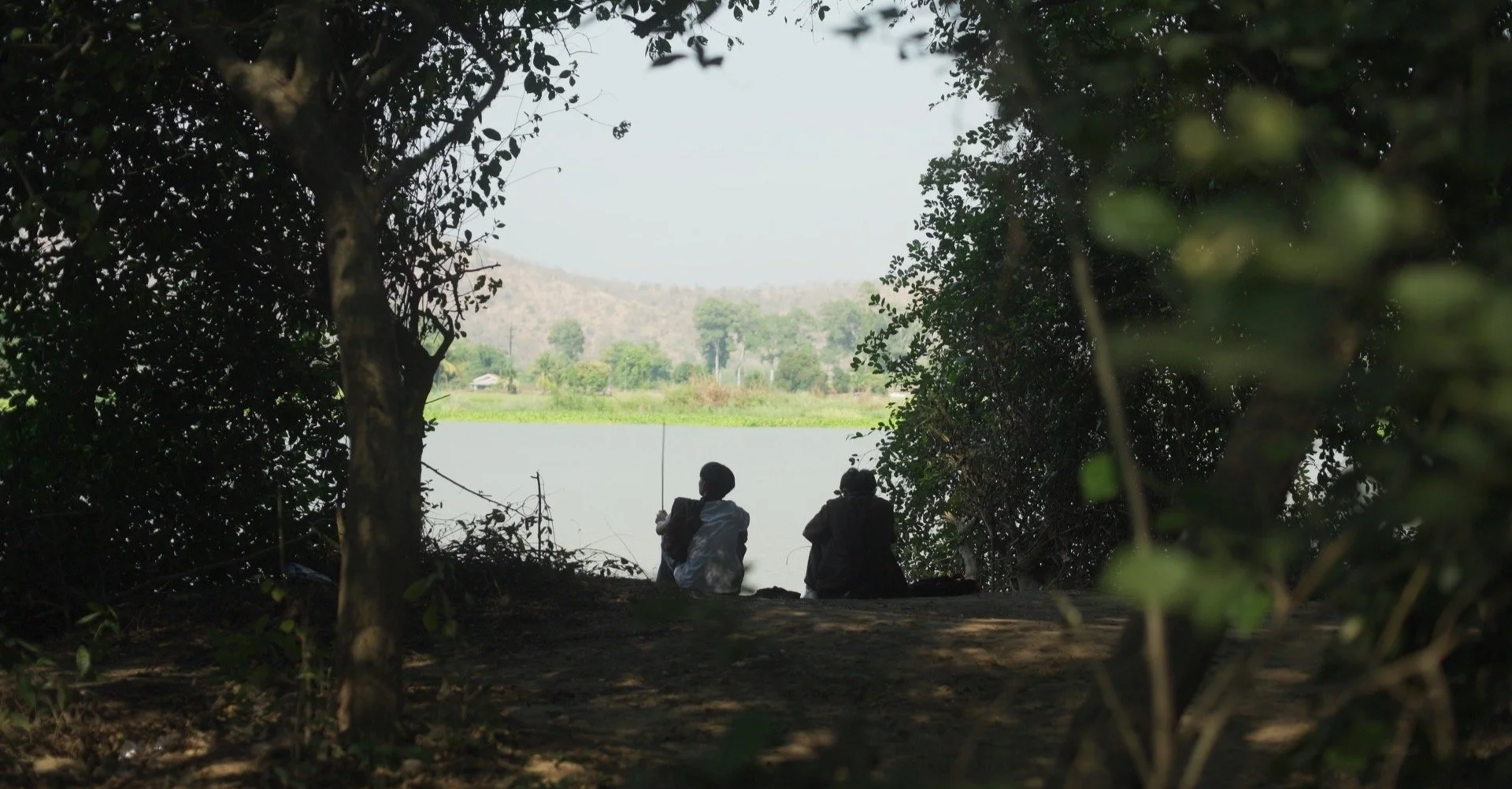 Two children sitting under trees near a body of water, with hills and greenery in the background, viewed from shade within a wooded area.
