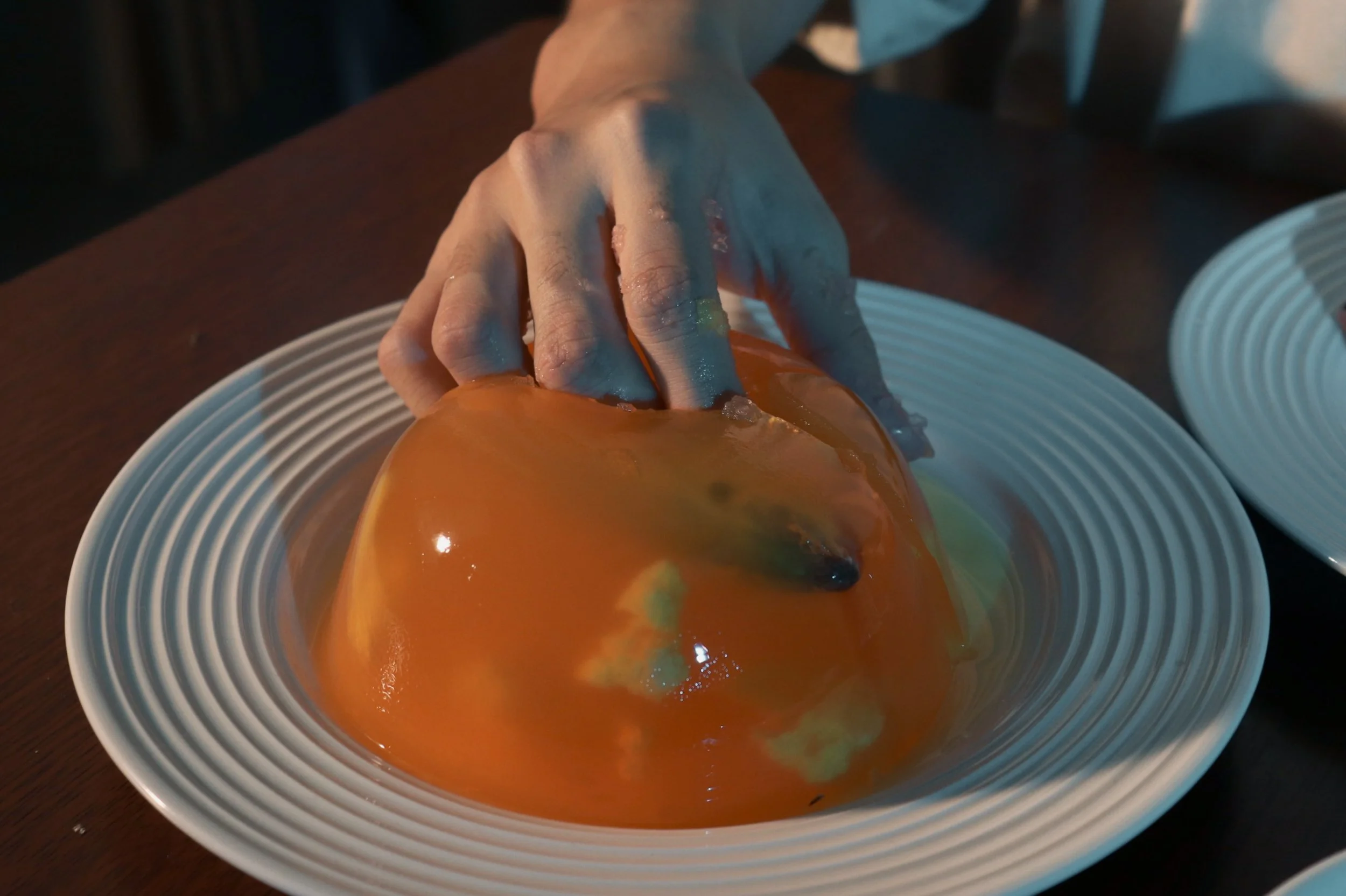 A person reaching into a bright orange Halloween pumpkin bowl with green and black patches, placed on a striped white plate. The person is wearing a white shirt, and the background includes a dark wooden table and napkins.