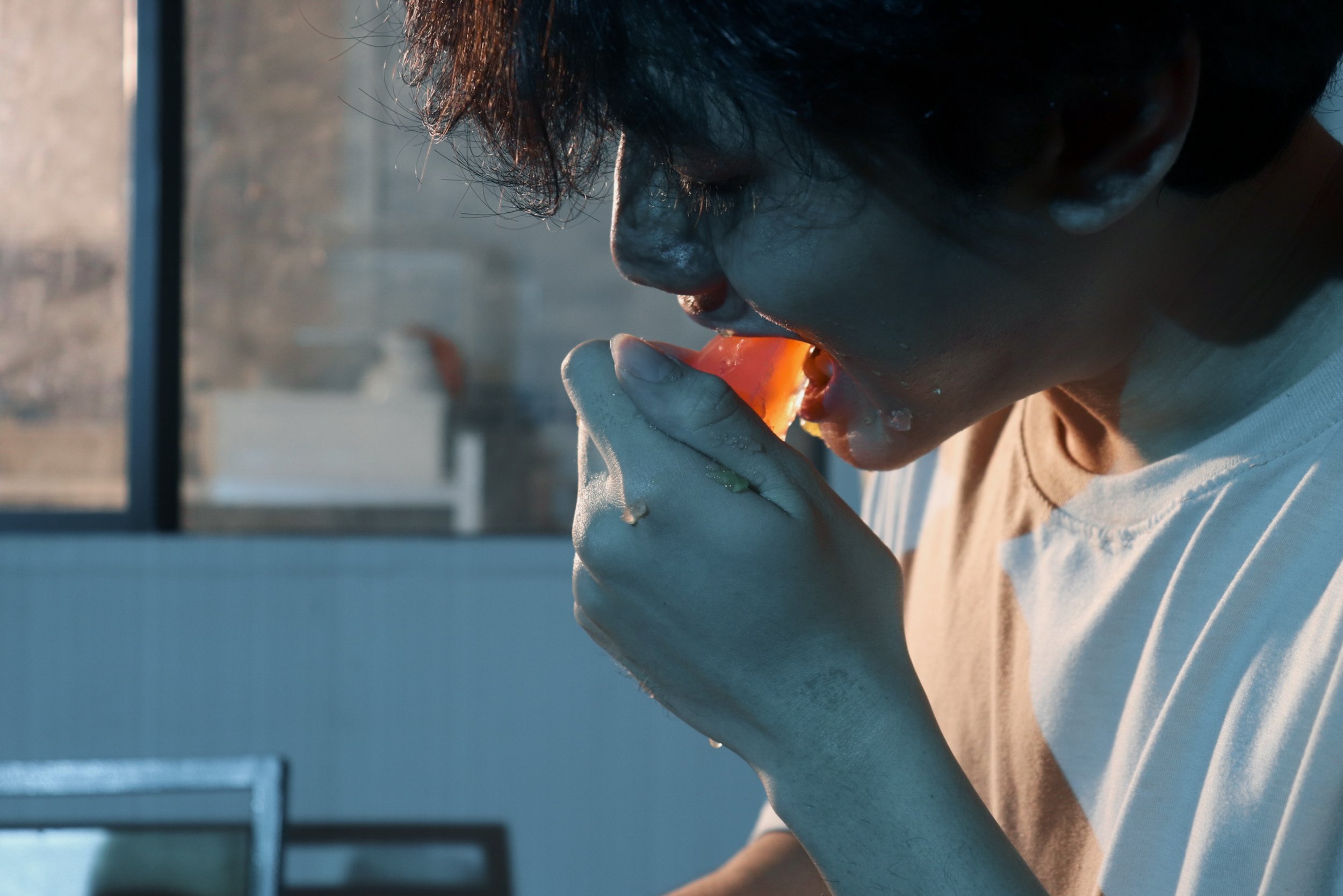 A person with dark, curly hair is about to take a bite of a slice of pizza, holding it with their hand. The scene is lit with natural sunlight and appears to be in a kitchen or dining area.