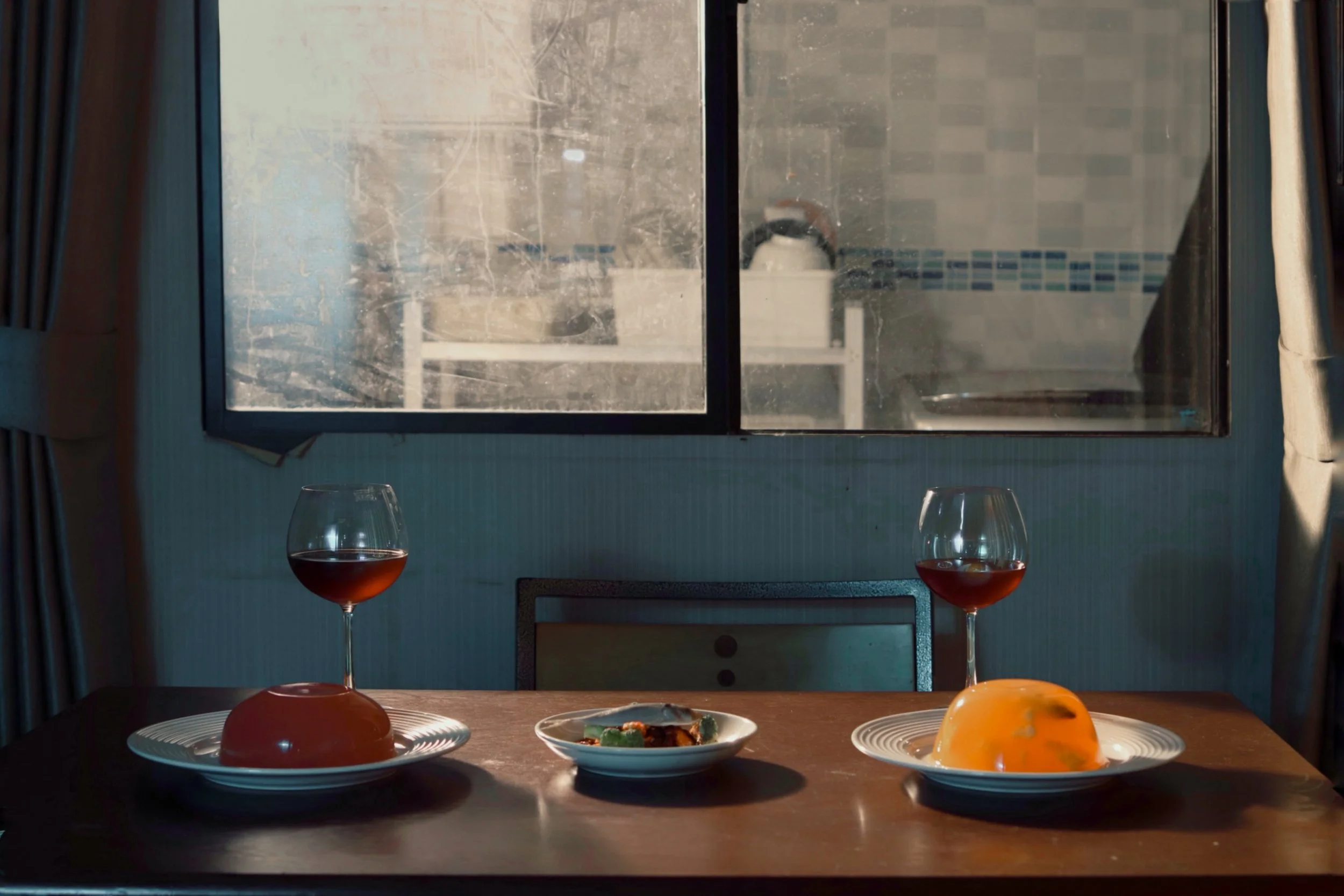 A dining table set with plates of food, including a dish with greens and meat, and glasses of red wine. In the background, a window with curtains and a view of a kitchen with a sink and tiled wall are visible.