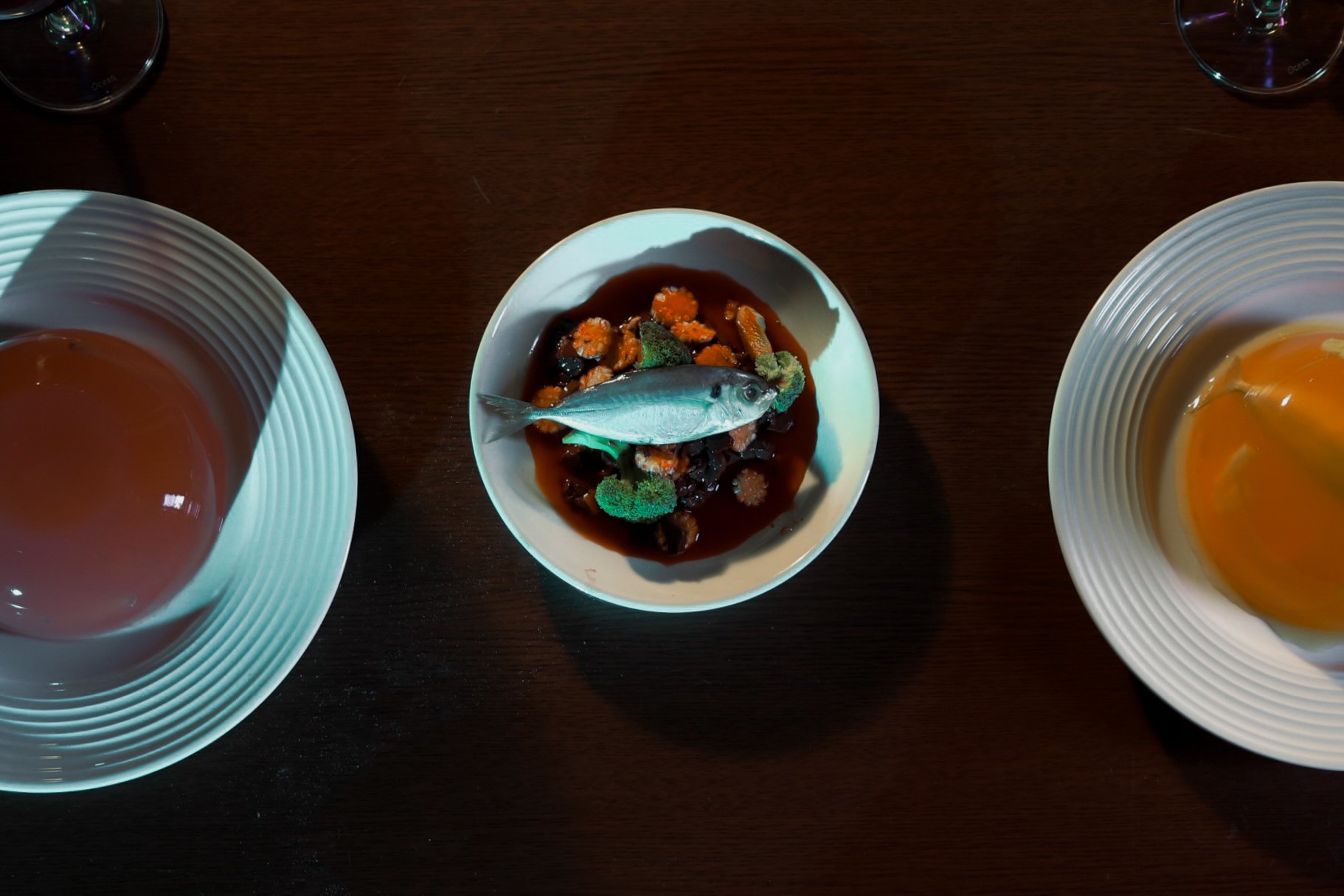 A seafood dish with a small fish on top of vegetables and sauce in a white bowl, flanked by two other bowls containing what appears to be soup or broth, on a dark brown table.