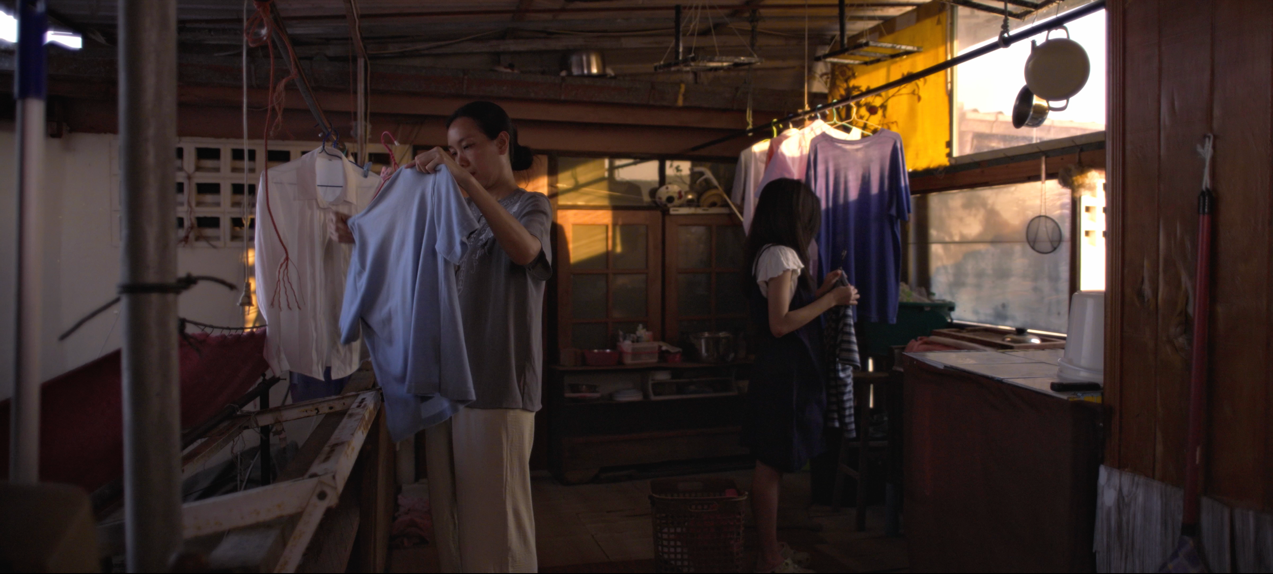 Two women in a rustic laundry room hanging clothes to dry, with sunlight streaming through a small window.