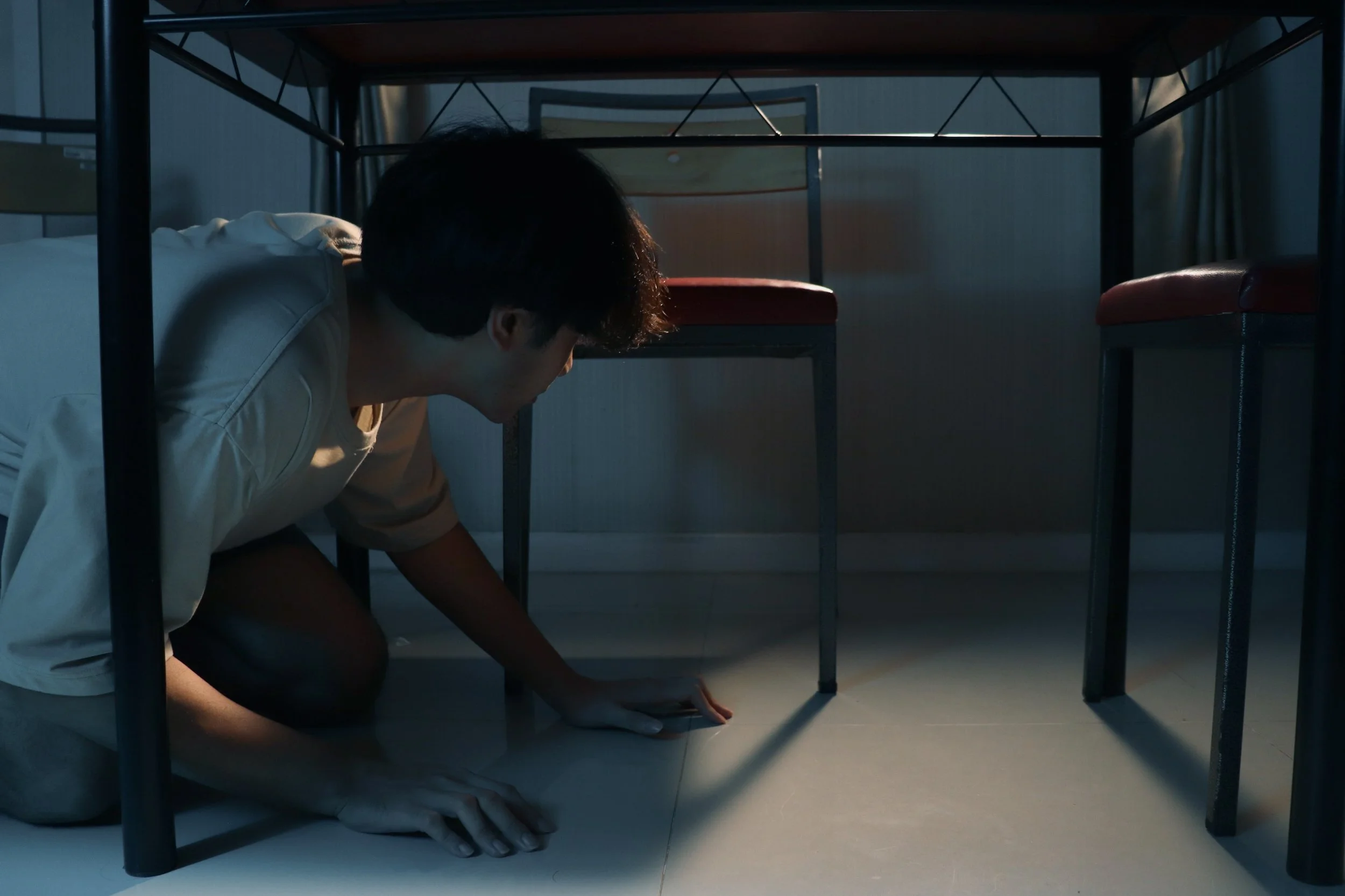 A person crouching under a table, reaching towards something on the floor, with chairs and a wall visible in the background.