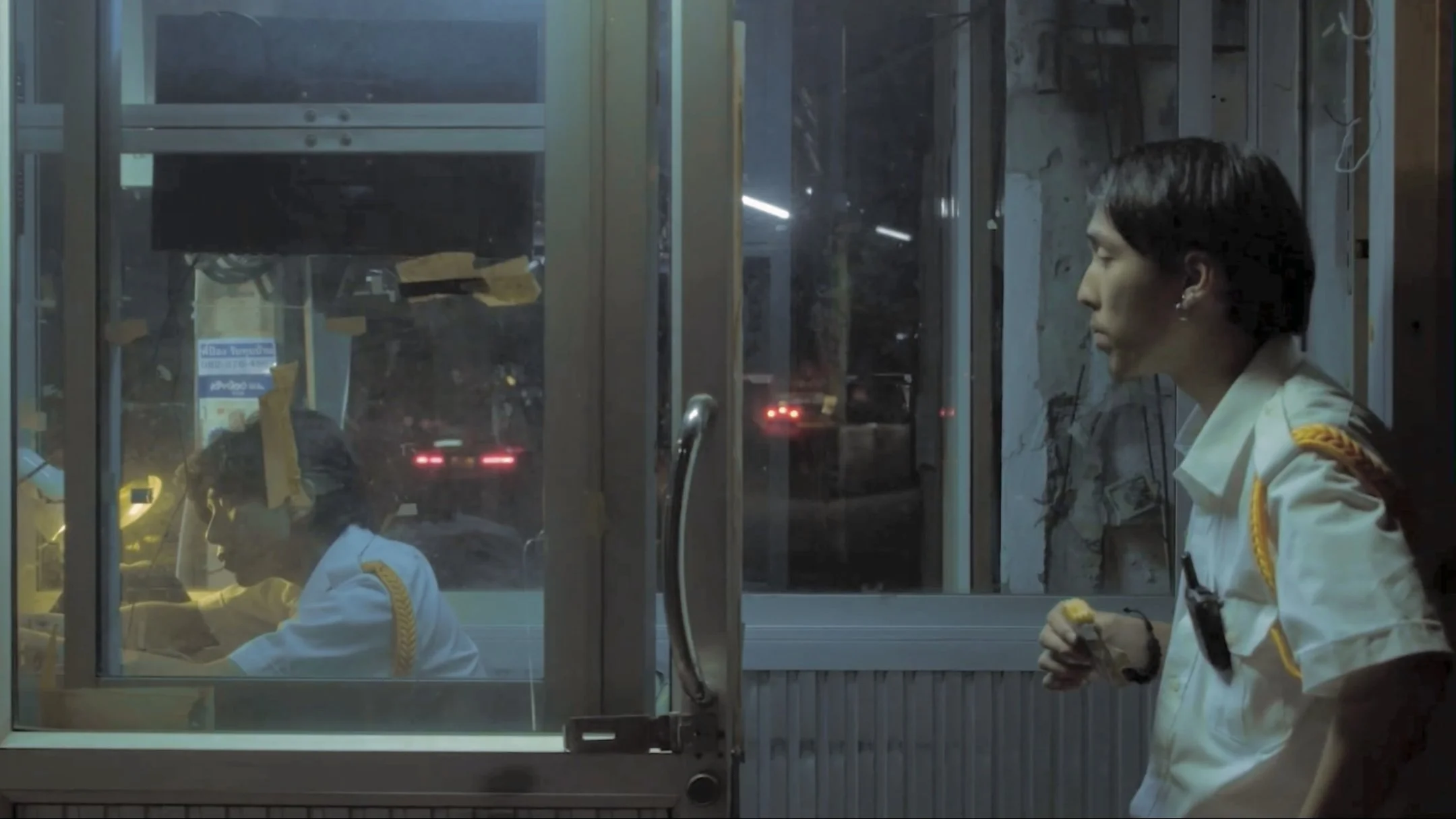 Two women wearing white uniforms and yellow shoulder cords inside a glass booth at night. One woman is seated and working, while the other stands holding a small object, possibly food. Outside the booth, cars are visible with taillights glowing in the darkness.