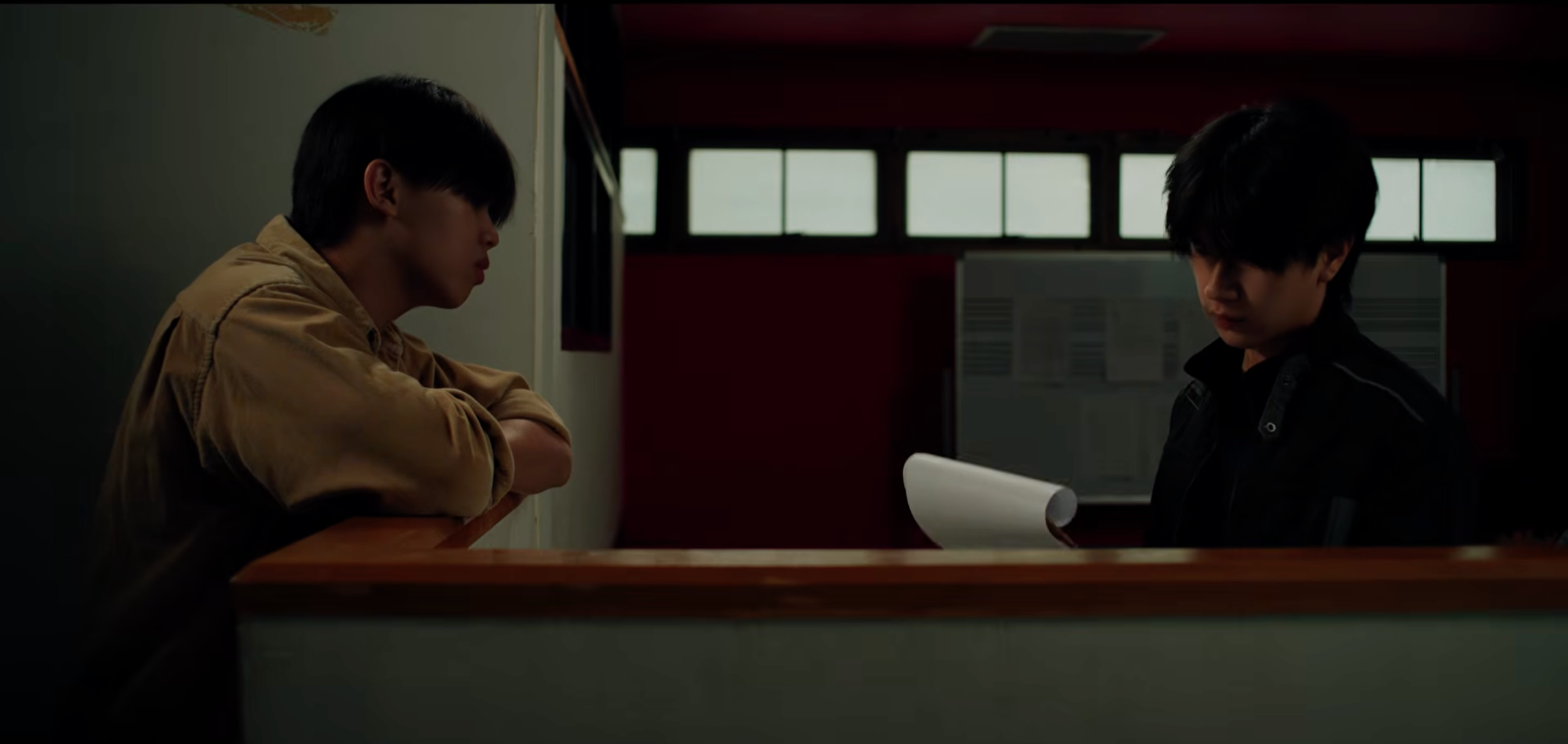 Two young men seated at a counter, engaged in conversation, in a dimly lit room with windows in the background.