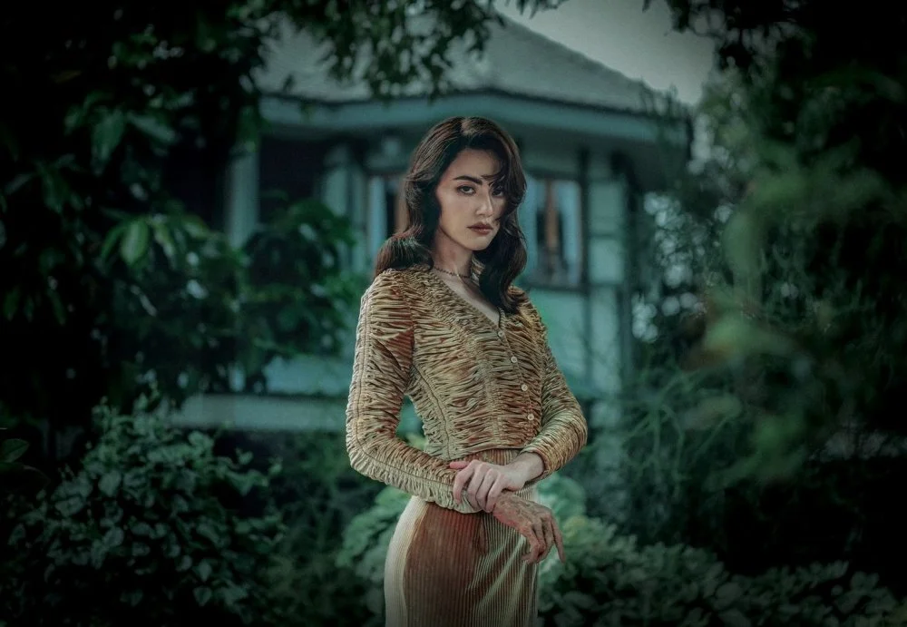 A young woman with dark hair in loose waves, wearing a textured gold dress, standing outdoors with trees and a house in the background.