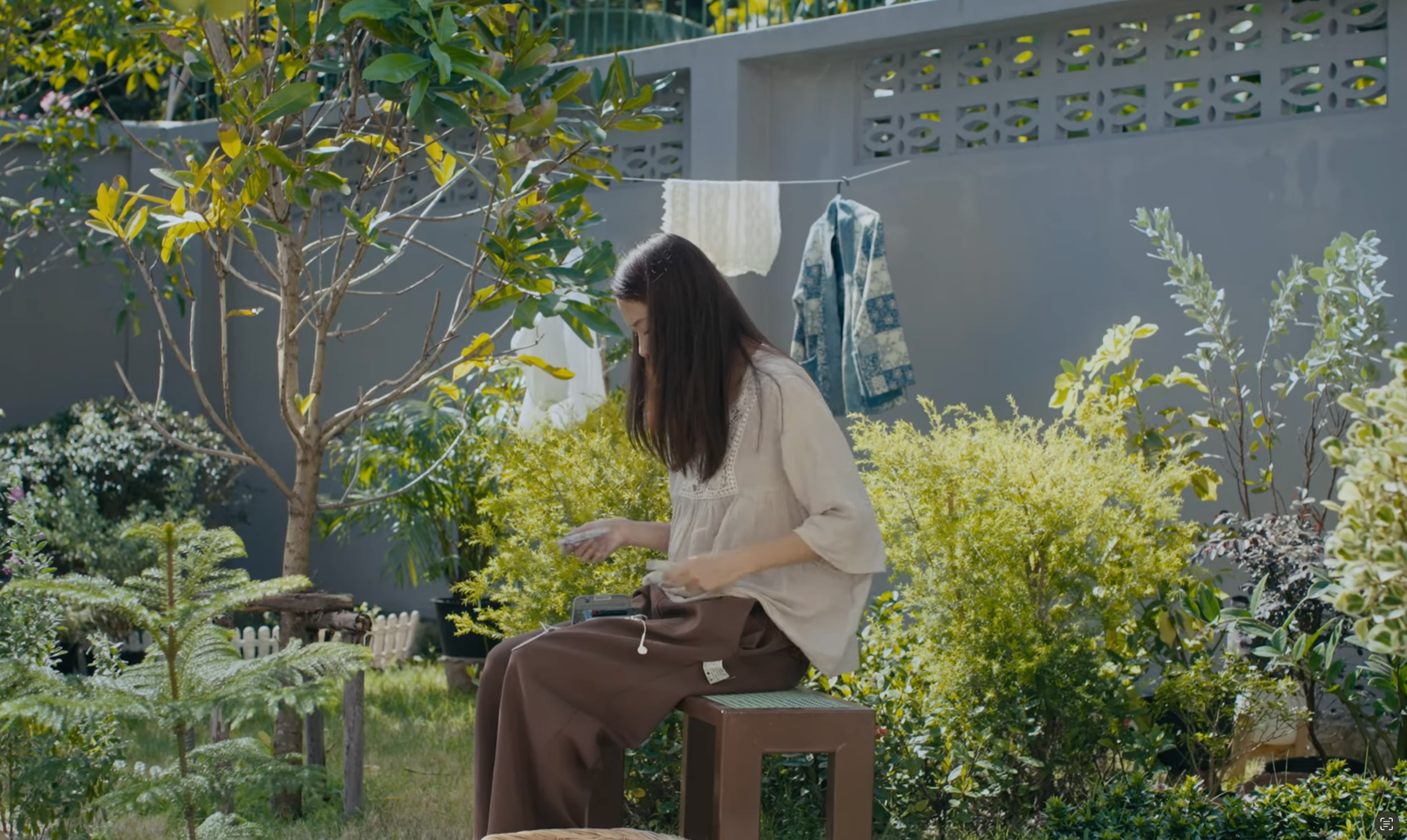 A woman with long dark hair sitting on a bench in a lush garden, looking down at something in her hands. There are green plants and trees around her, with laundry hanging on a line behind her and a gray wall with decorative cutouts in the background.