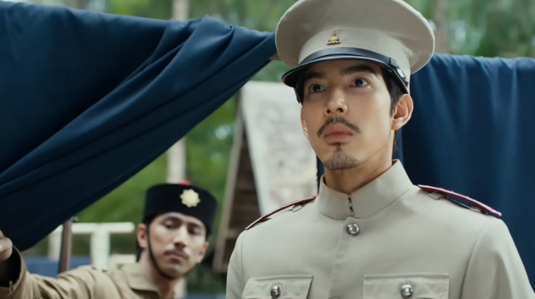 A man in a beige military uniform with a white hat salutes during a formal ceremony, with other uniformed individuals in the background.
