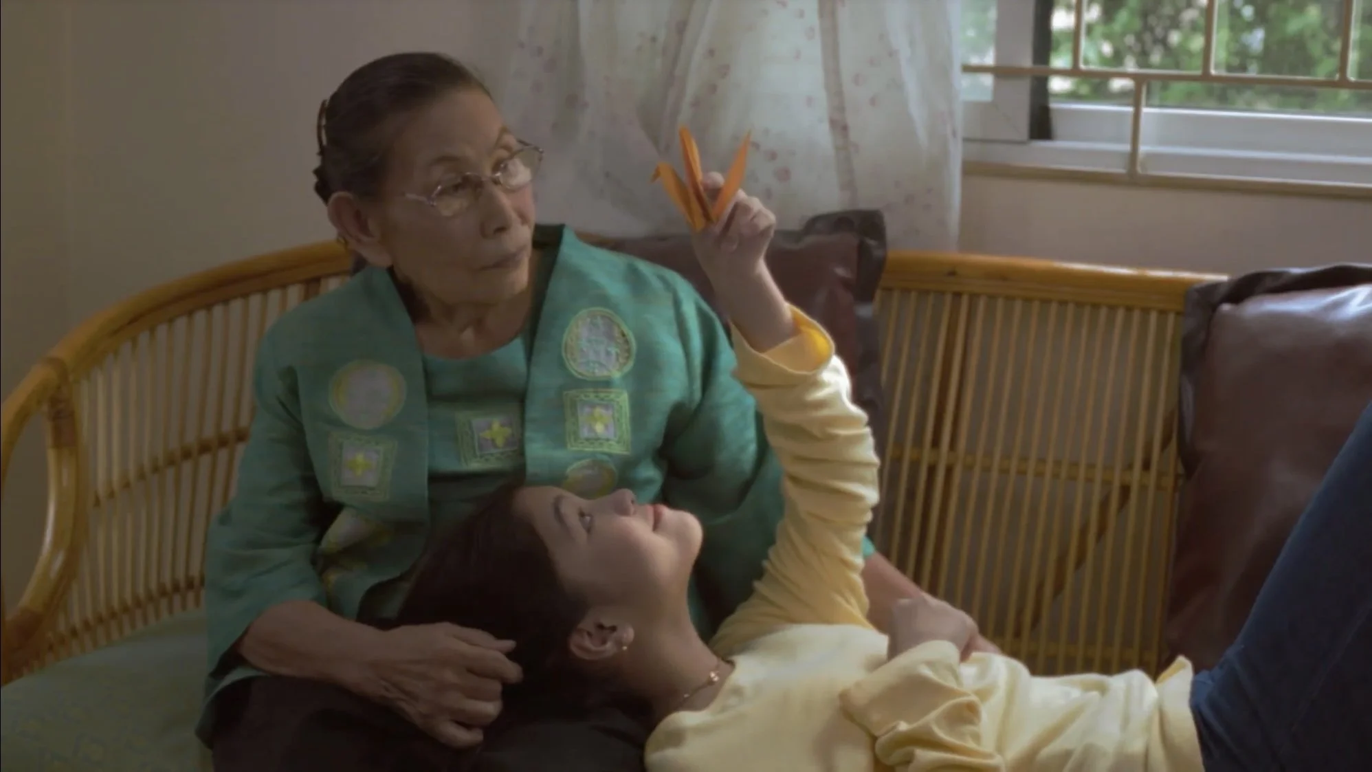 An elderly woman wearing glasses and a turquoise top with embroidered patches, sitting on a bed and holding a handful of orange pencils, with a young girl lying on her lap, looking up at her. They are in a well-lit room with a window and curtain in the background.