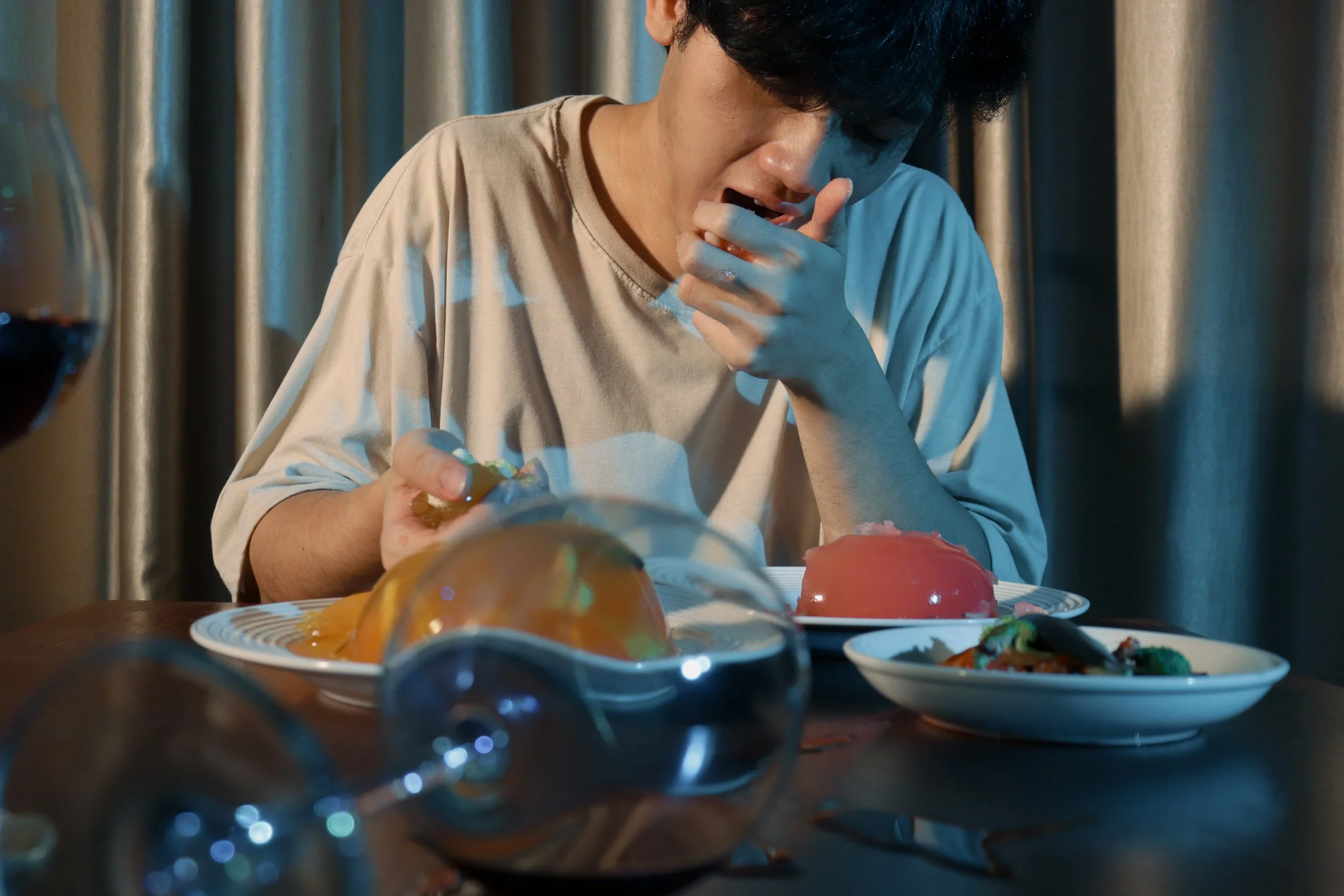 A person with dark hair wearing a beige shirt, sitting at a table filled with various colorful desserts, appears to be tasting or smelling a dessert with their hand near their mouth. The background features a curtain.