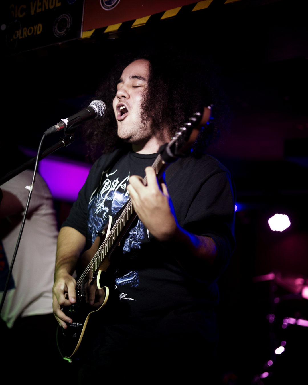 A male musician with curly hair singing and playing an electric guitar on stage during a live performance.