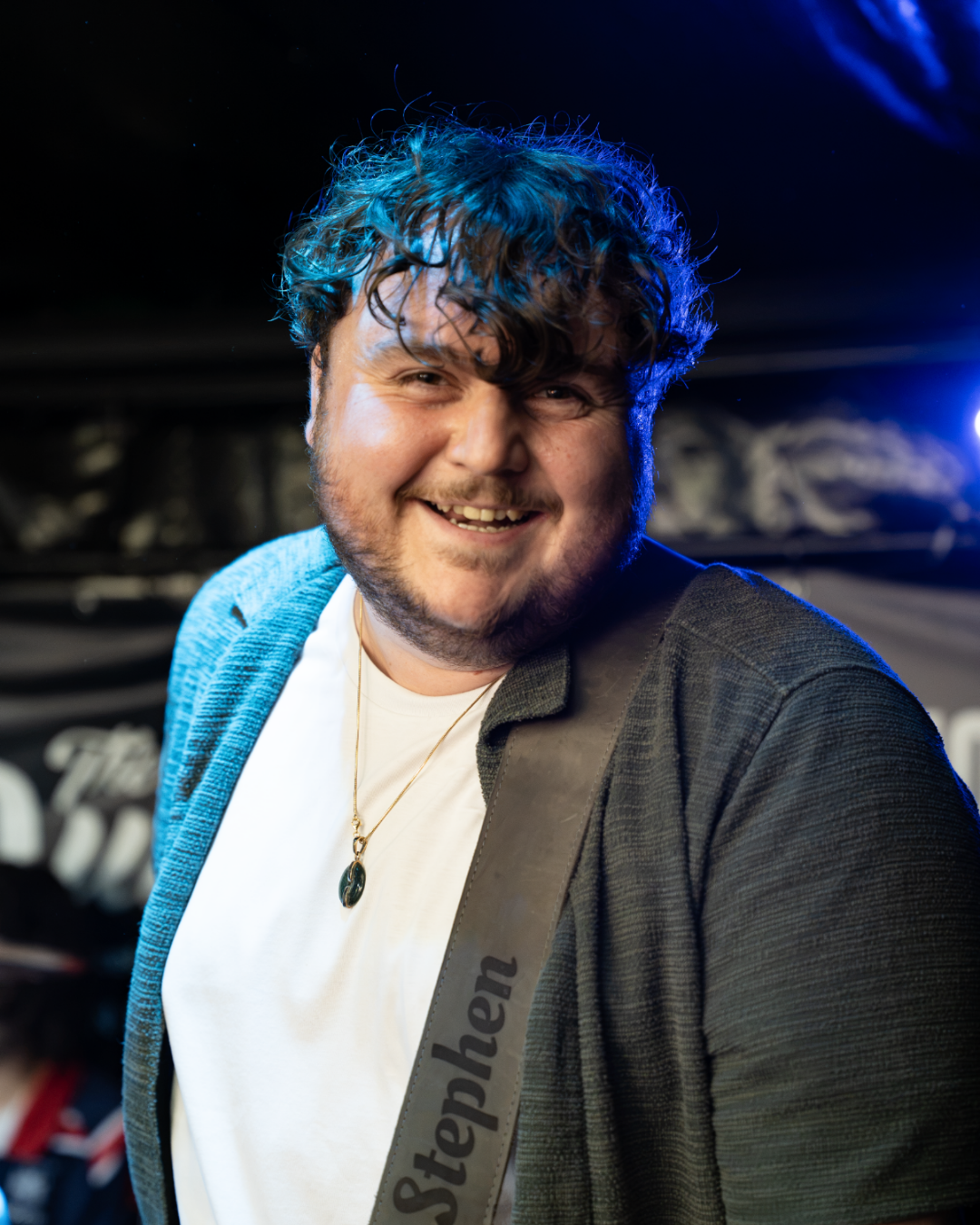 Smiling young man with wavy dark hair, wearing a white t-shirt, a dark jacket, a necklace, and a guitar strap with the name 'Stephen' on it, in a dimly lit indoor setting.