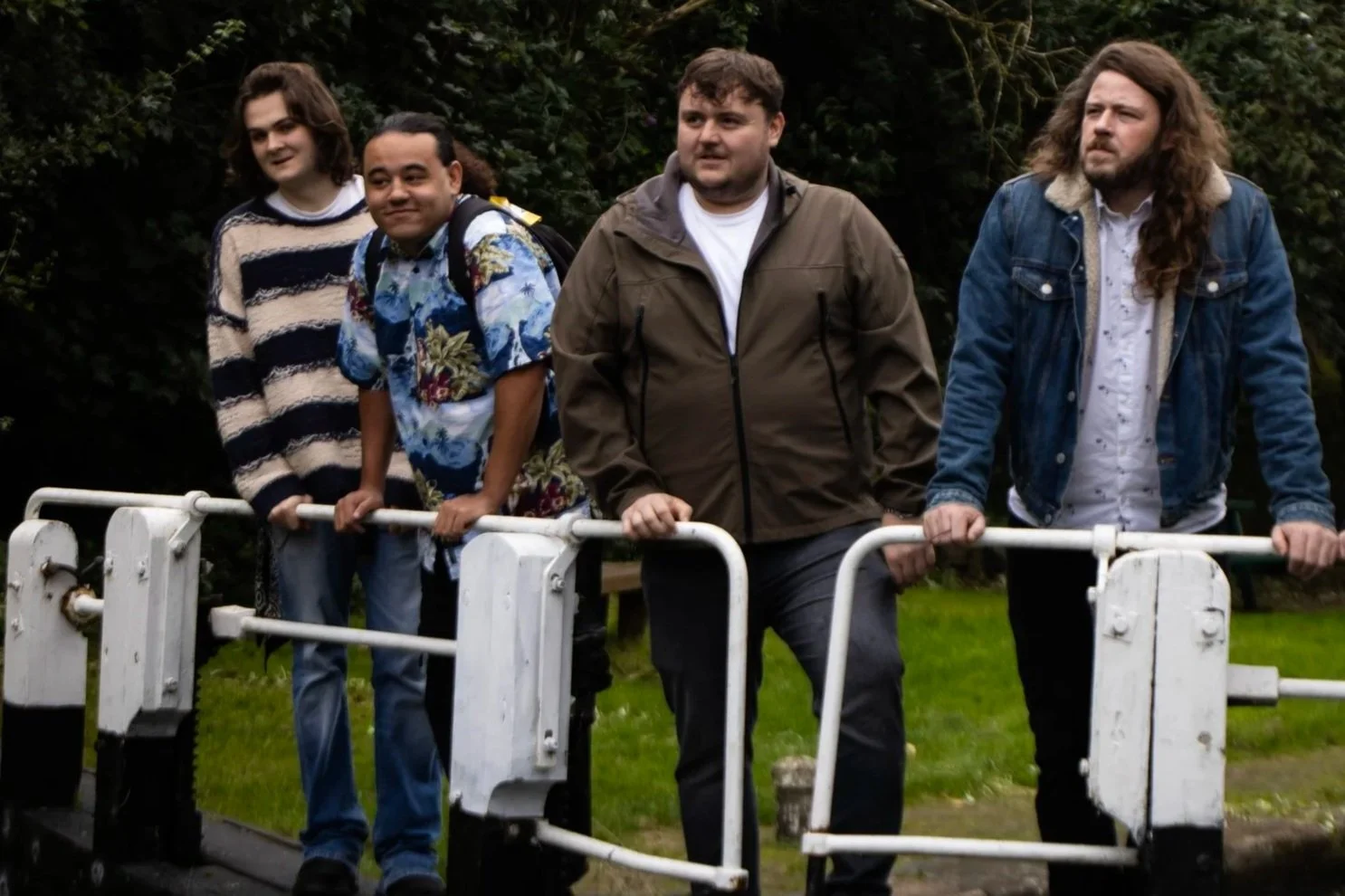 Four men standing outdoors behind a white metal barrier, with trees and greenery in the background.