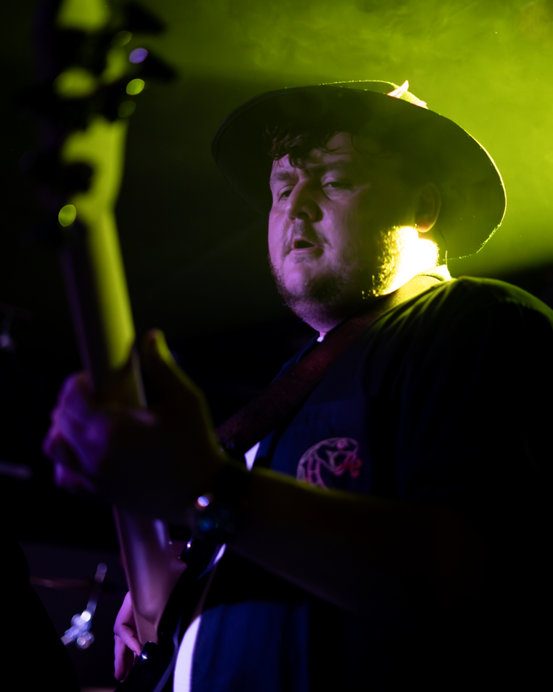 A man playing electric guitar on stage, wearing a black hat and dark clothing, illuminated by green stage lighting.