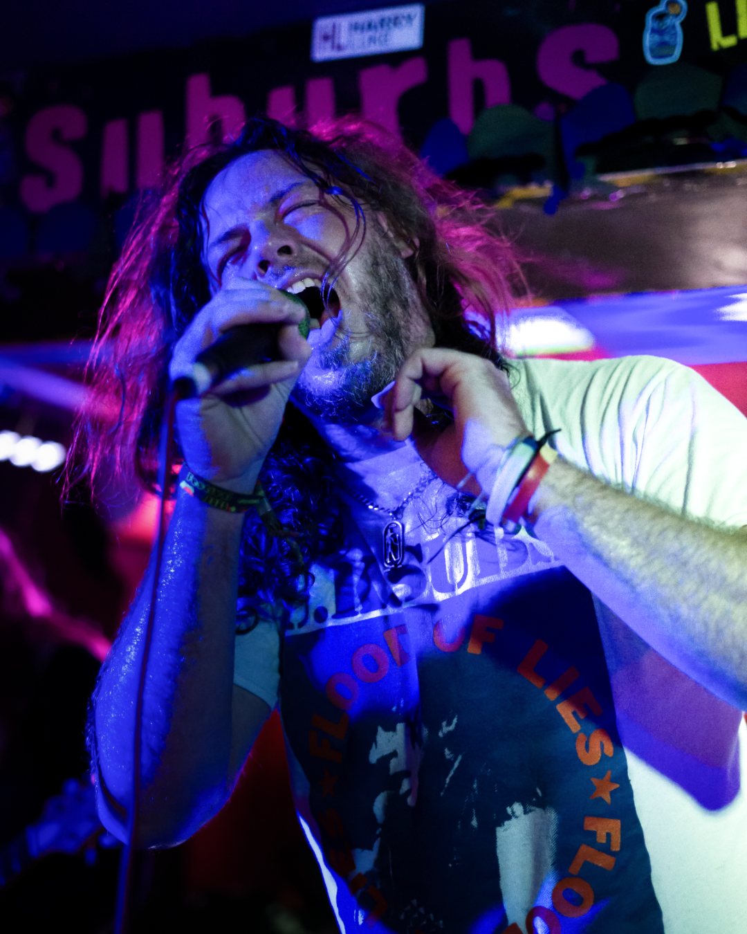 A male singer with long, curly hair passionately singing into a microphone during a live concert, with purple and blue stage lighting.