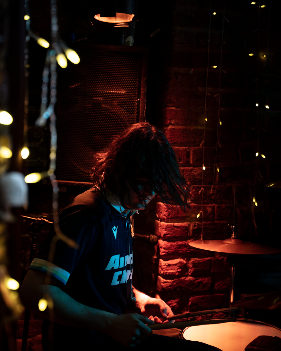 A person with long hair, wearing a dark jersey, sitting in a dimly lit space and playing a drum.