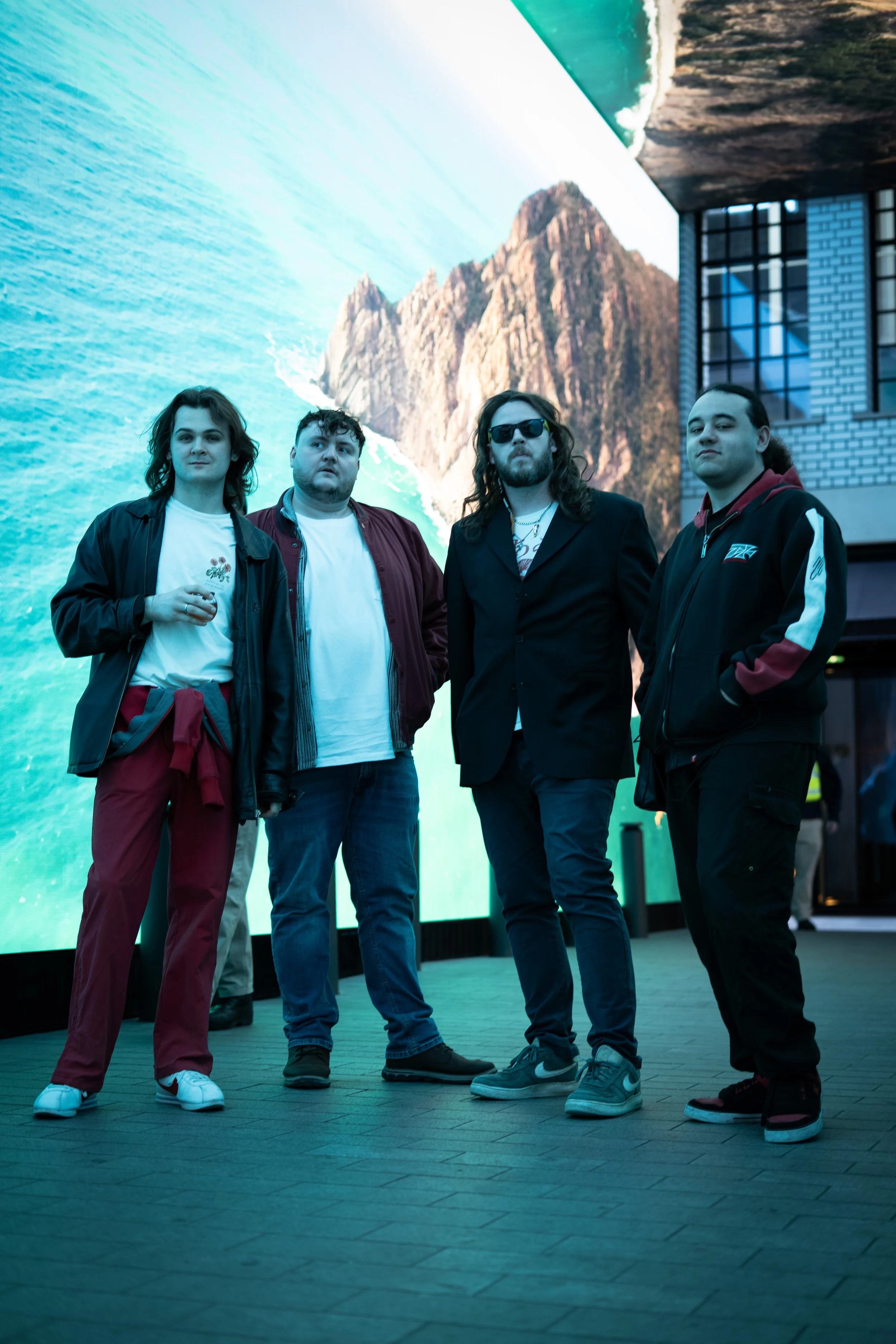 Four young men standing in front of a large outdoor digital screen displaying a mountain landscape with ocean waves. They are dressed casually, some with sunglasses and jackets, and are posing for the photo.