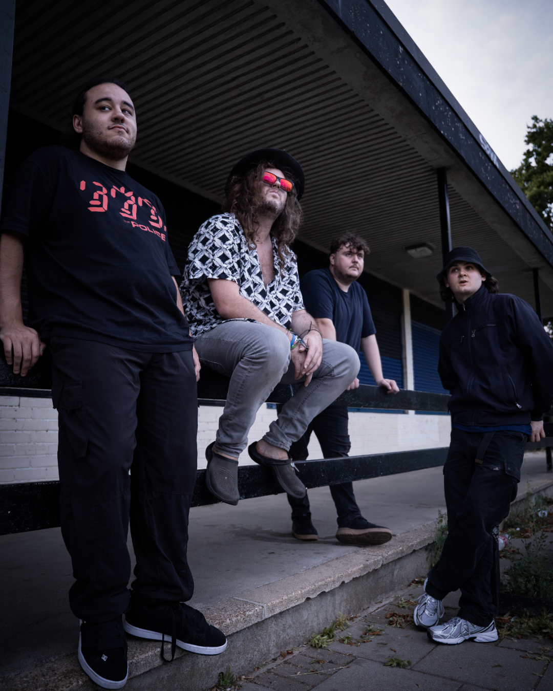 Four young men outside near a building, with one sitting on a railing, wearing sunglasses and a patterned shirt, and the others standing around, dressed casually.