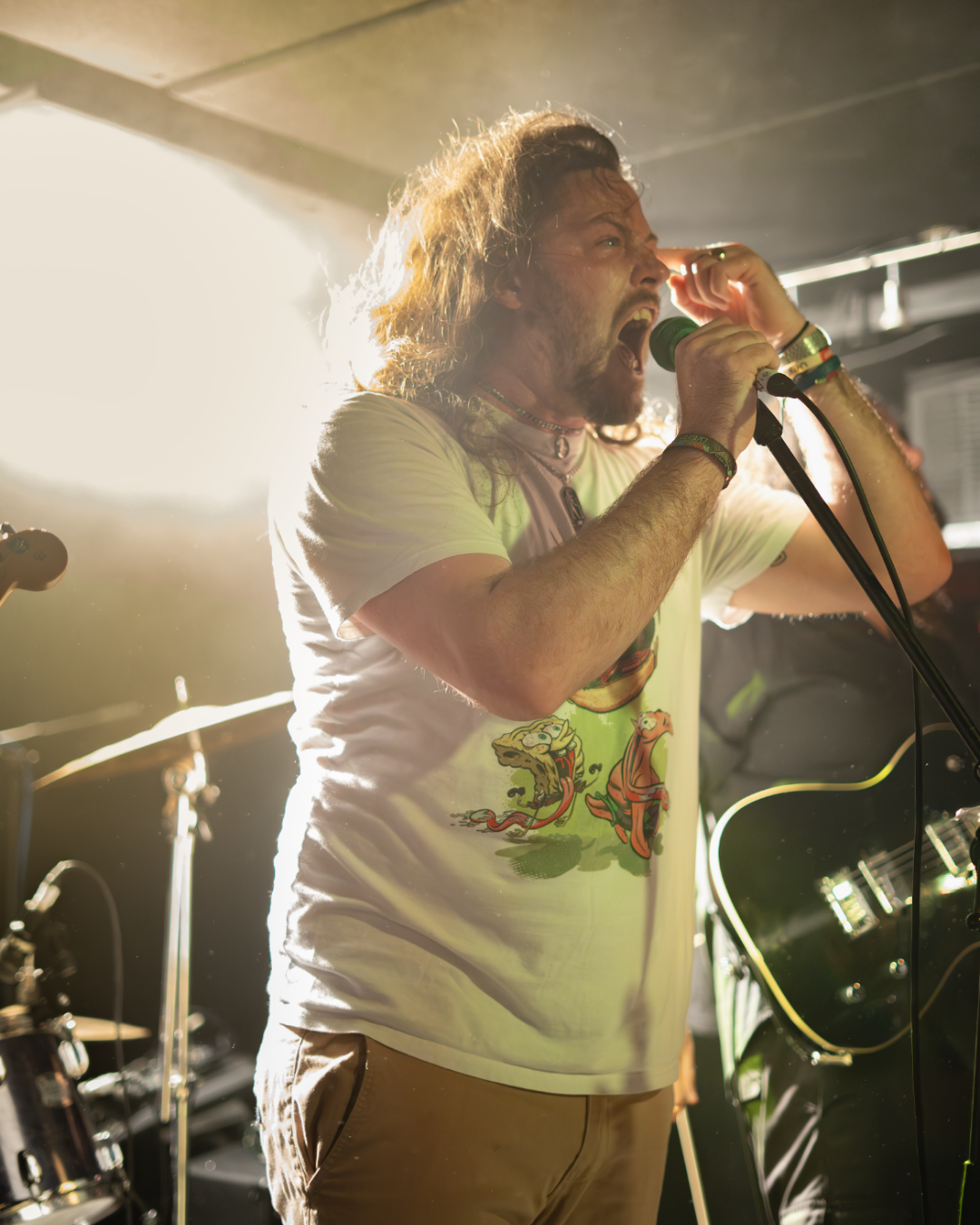 Male singer with long hair and beard singing into a microphone, wearing a graphic t-shirt, on stage with bright backlighting and musical equipment in the background.