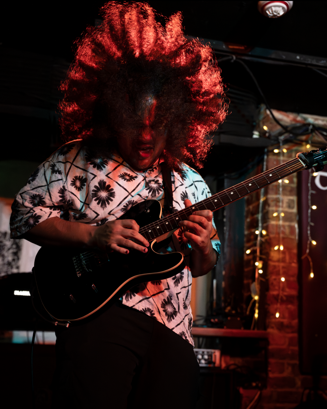 A person with voluminous curly hair playing an electric guitar on stage, wearing a patterned shirt, with stage lights and string lights in the background.
