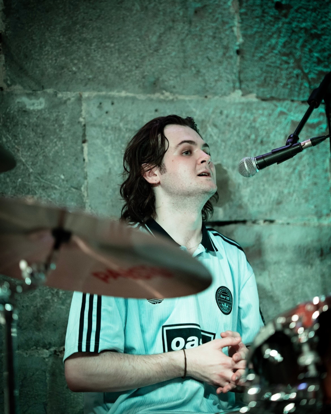 A young man with long dark hair singing or speaking into a microphone while sitting behind a drum set against a stone wall background.